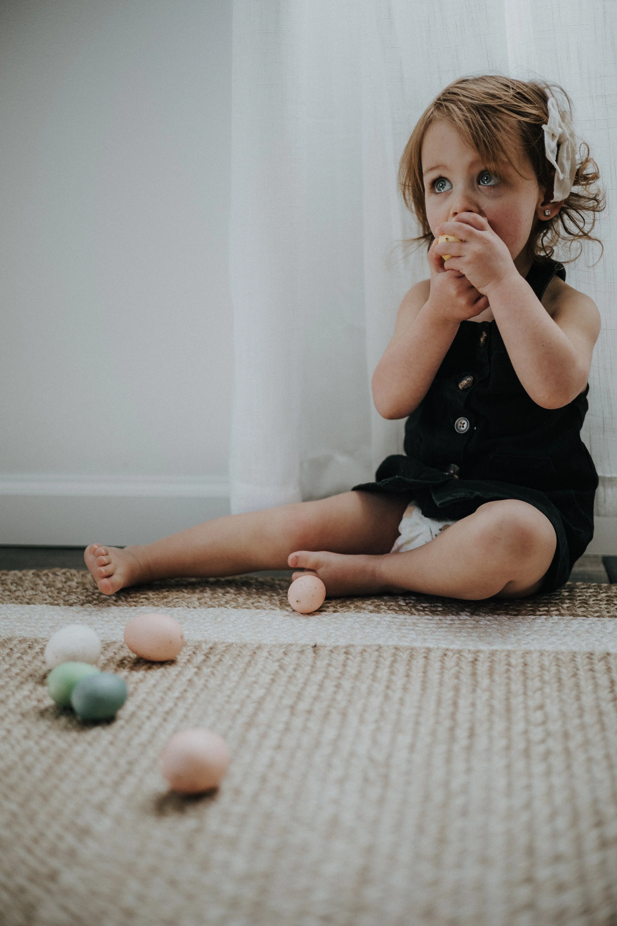 A young girl with curly red hair and blue eyes sitting on a woven rug, eating a snack, with colorful eggs scattered around her for easter photography in ohio