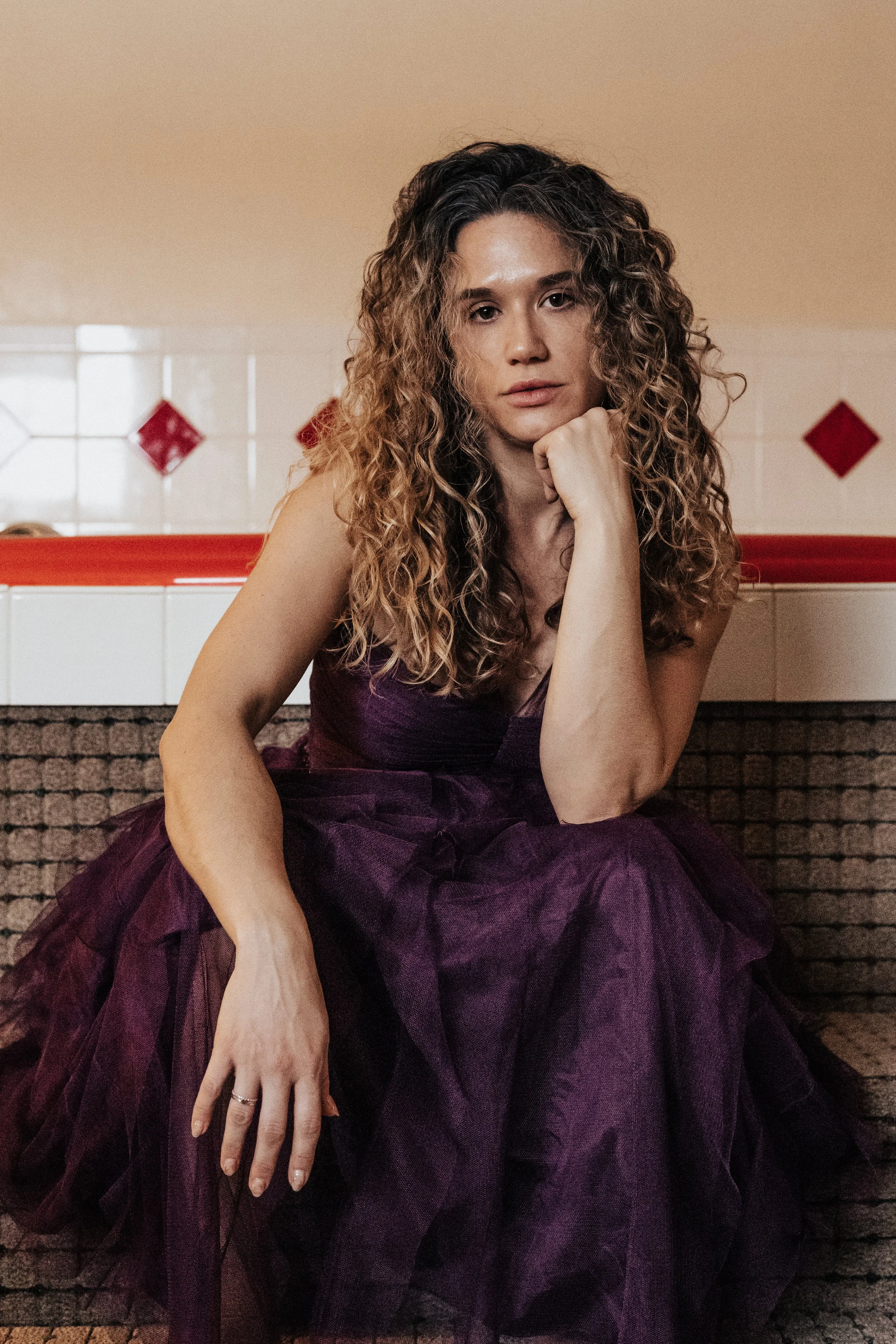 Woman with curly hair in a purple dress sitting on a bench in front of a tiled wall with red diamond accents in a vintage roadside motel in ohio