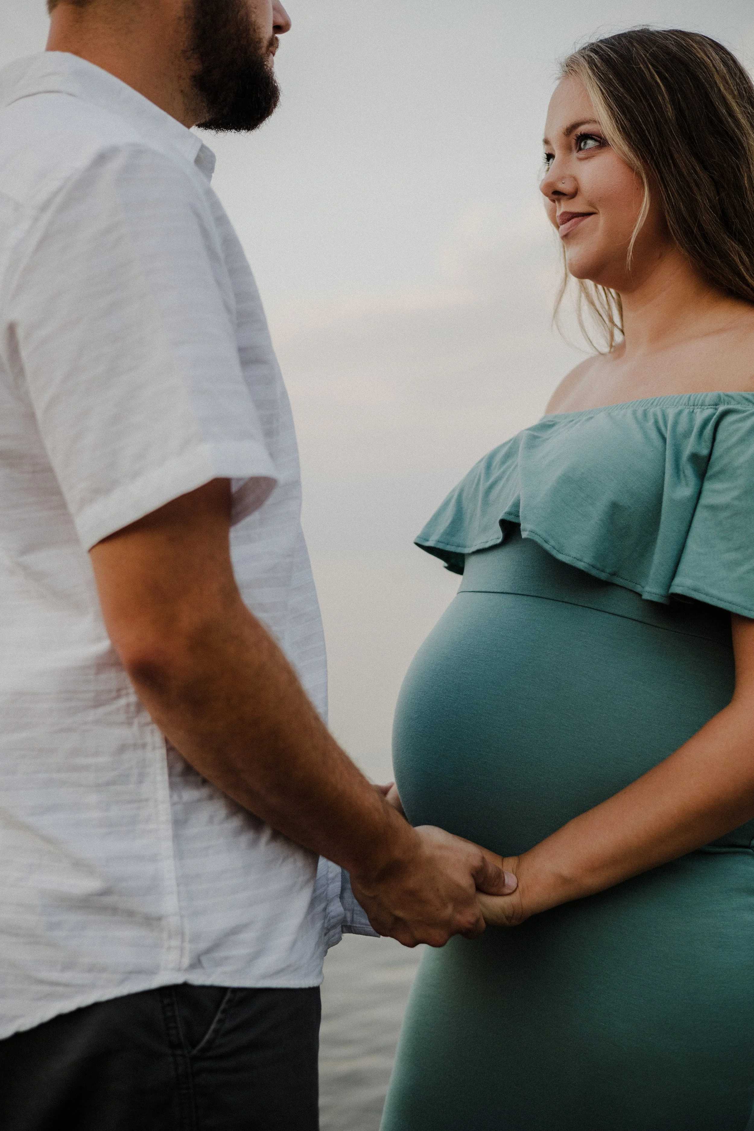 A pregnant woman and a man holding hands outdoors near water, facing each other, with the woman smiling.