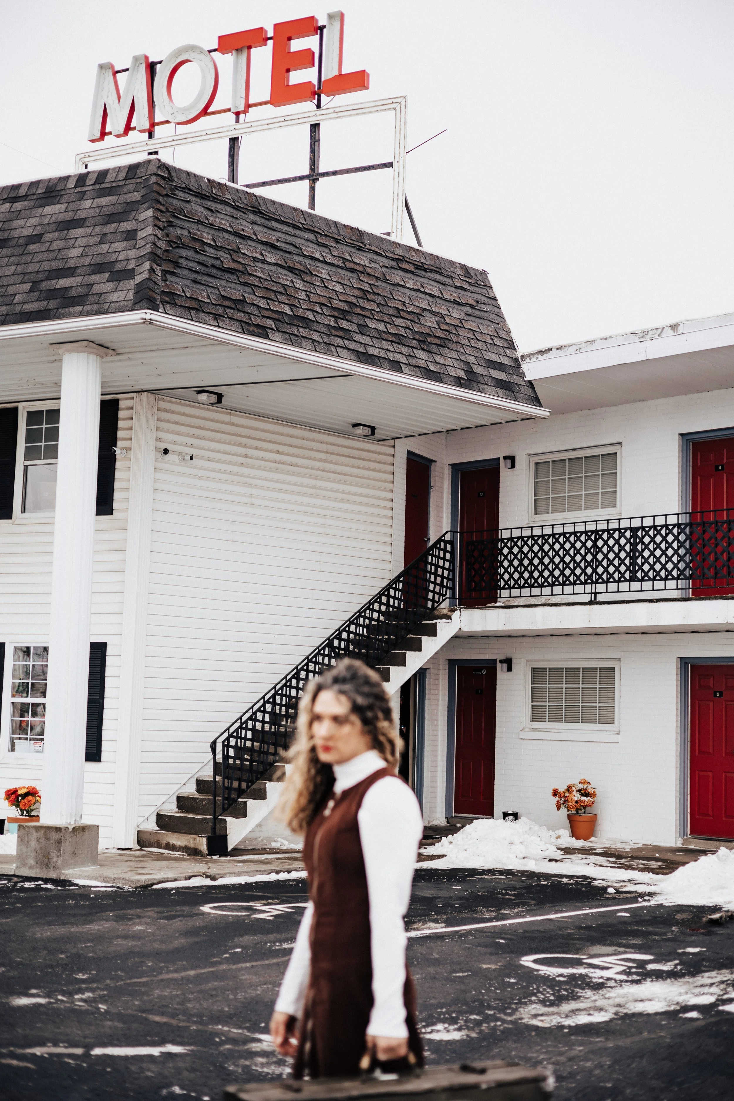 A woman with curly hair standing in front of a motel with white walls, red doors, and a black staircase, with a sign that reads "MOTEL" on the roof and snow on the ground.