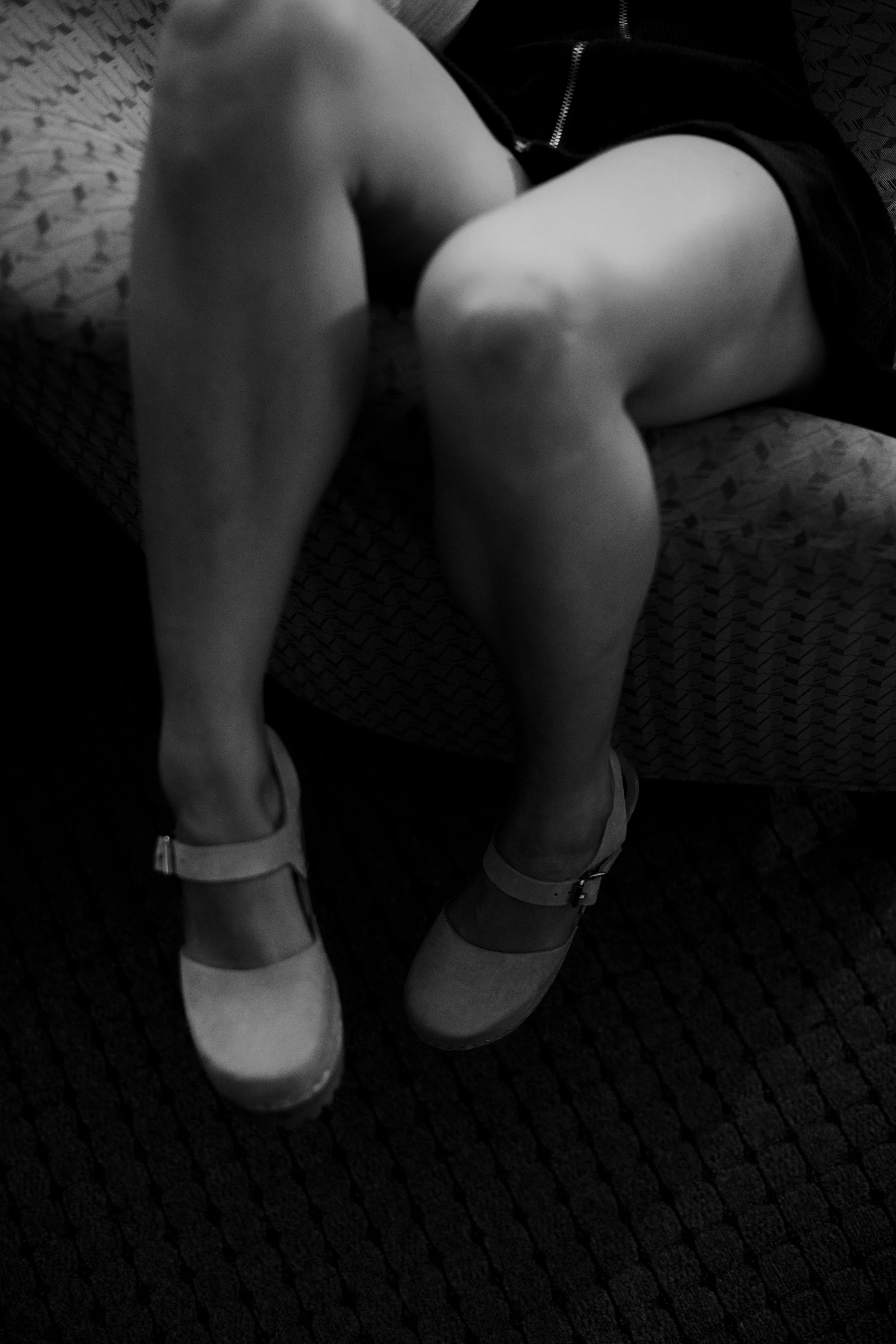 Person sitting on a patterned chair with legs bent and feet resting on the floor, wearing shoes.