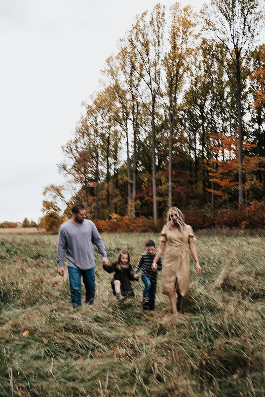 A family of four walking through a grassy field surrounded by tall trees with autumn foliage.