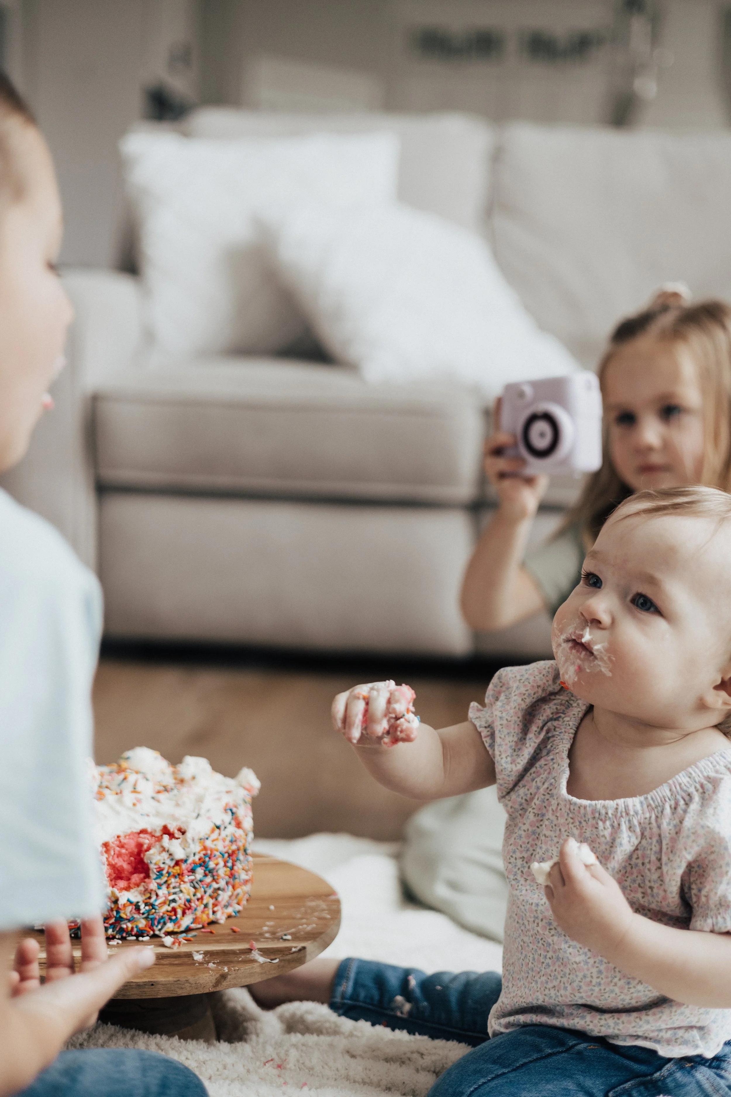 A young girl taking a photo of a younger child celebrating a birthday with a cake covered in rainbow sprinkles at an in home family photography session in monroeville ohio