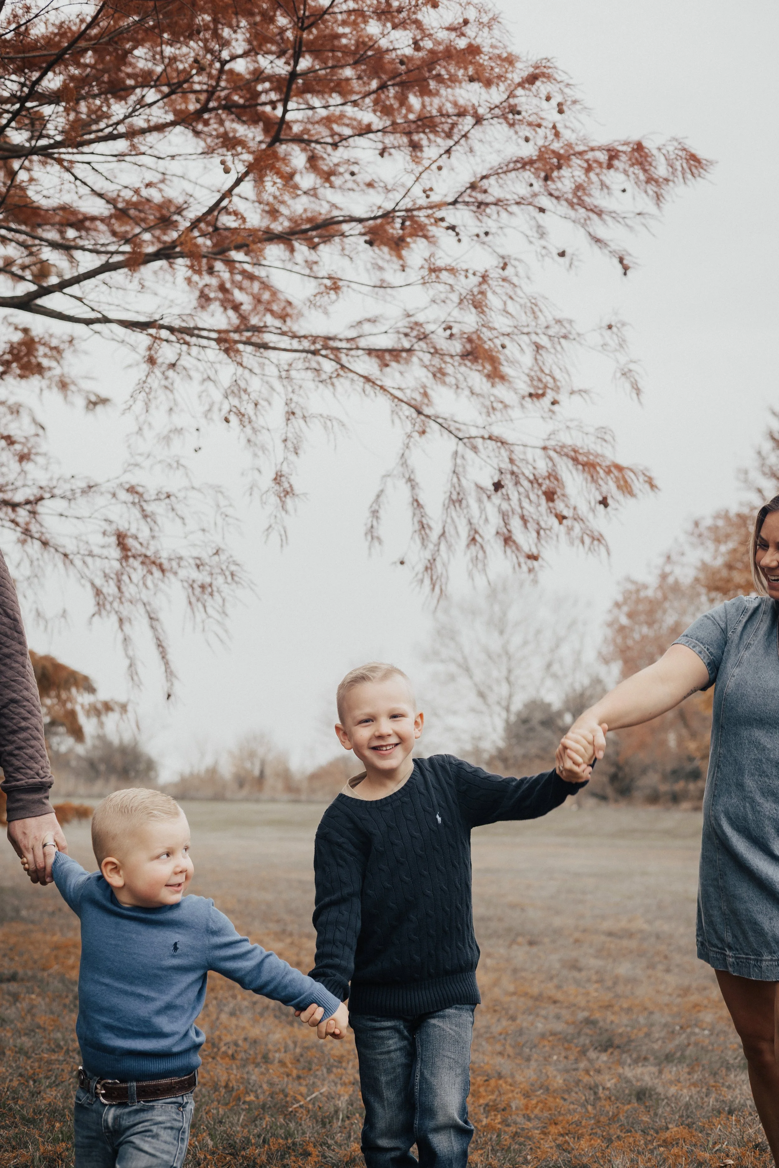 A smiling boy and a younger child holding hands and walking outdoors in fall, with a woman holding the older boy's hand during their family lifestyle photography photoshoot in huron ohio