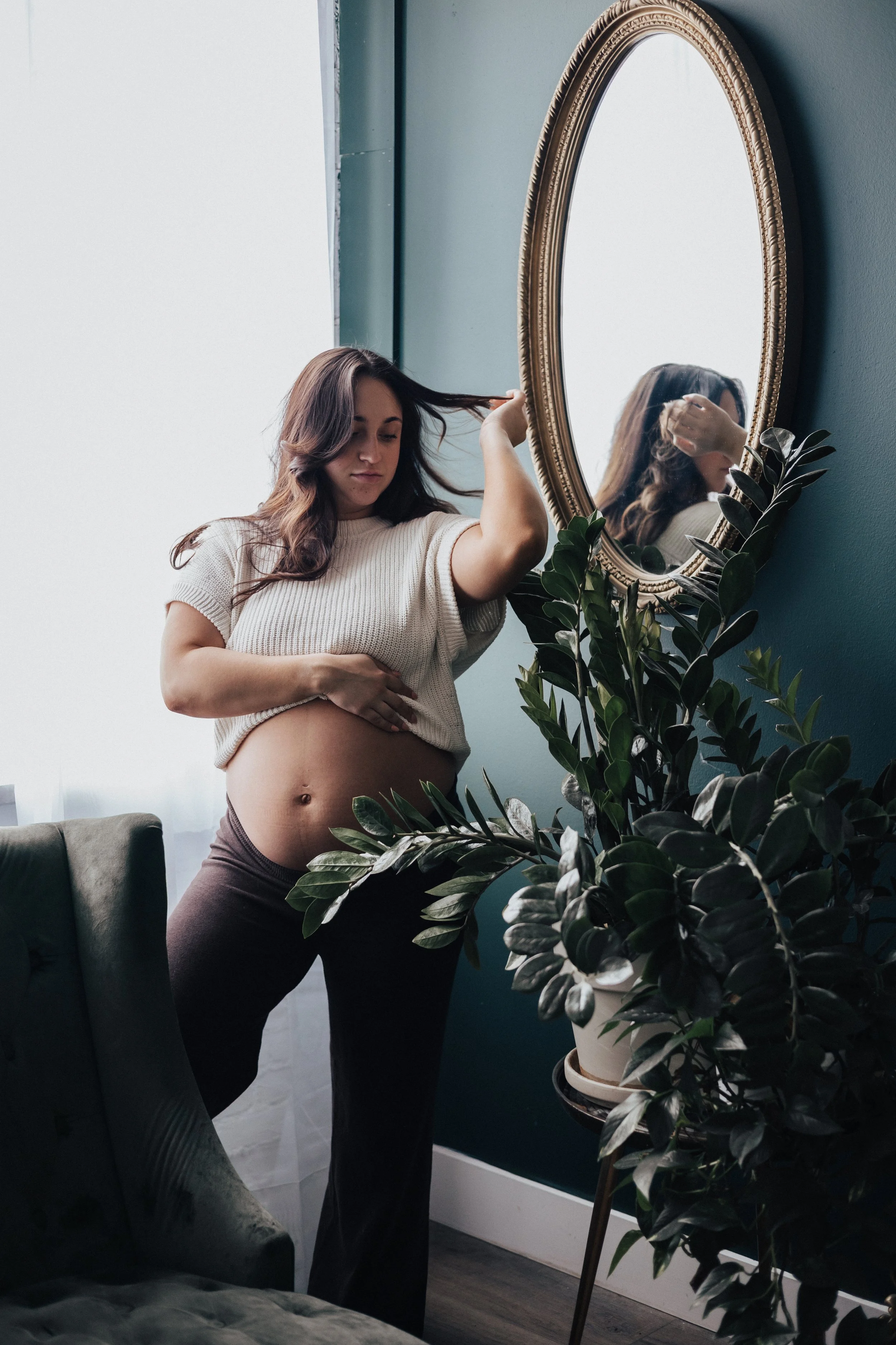 A pregnant woman standing in front of a mirror, touching her hair, while looking down at her belly. The room has a teal wall, a large oval mirror, a green potted plant, and a green armchair.