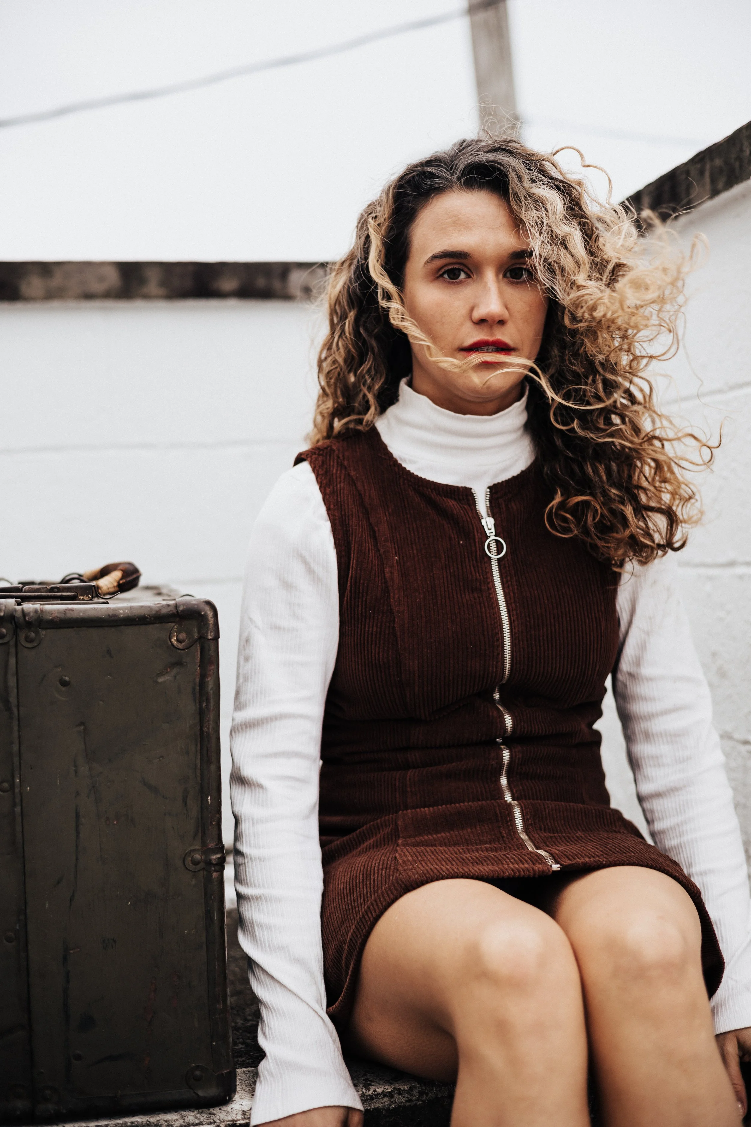 A young woman with curly hair, wearing a white turtleneck and a maroon corduroy dress, sitting outdoors next to a vintage suitcase at a roadside motel branding shoot in Avery ohio