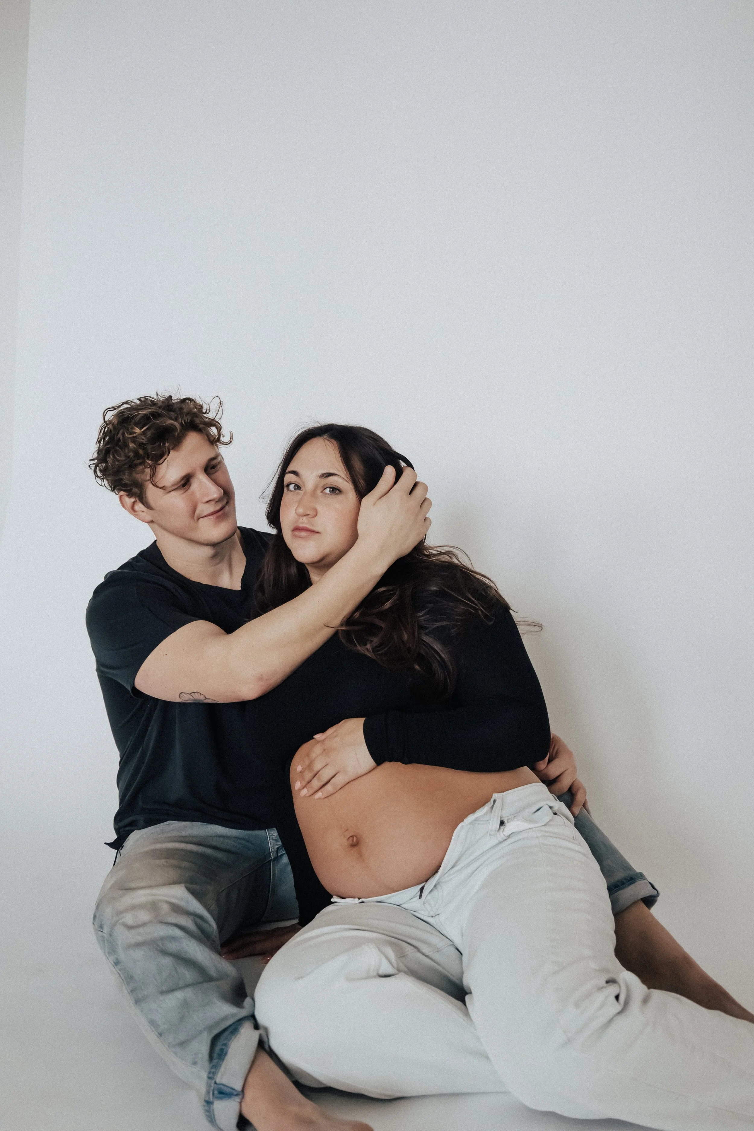 A couple sitting on the floor against a plain white wall, with the woman visibly pregnant, and the man gently holding her head and arm. pregnancy studio photography in Cleveland Ohio