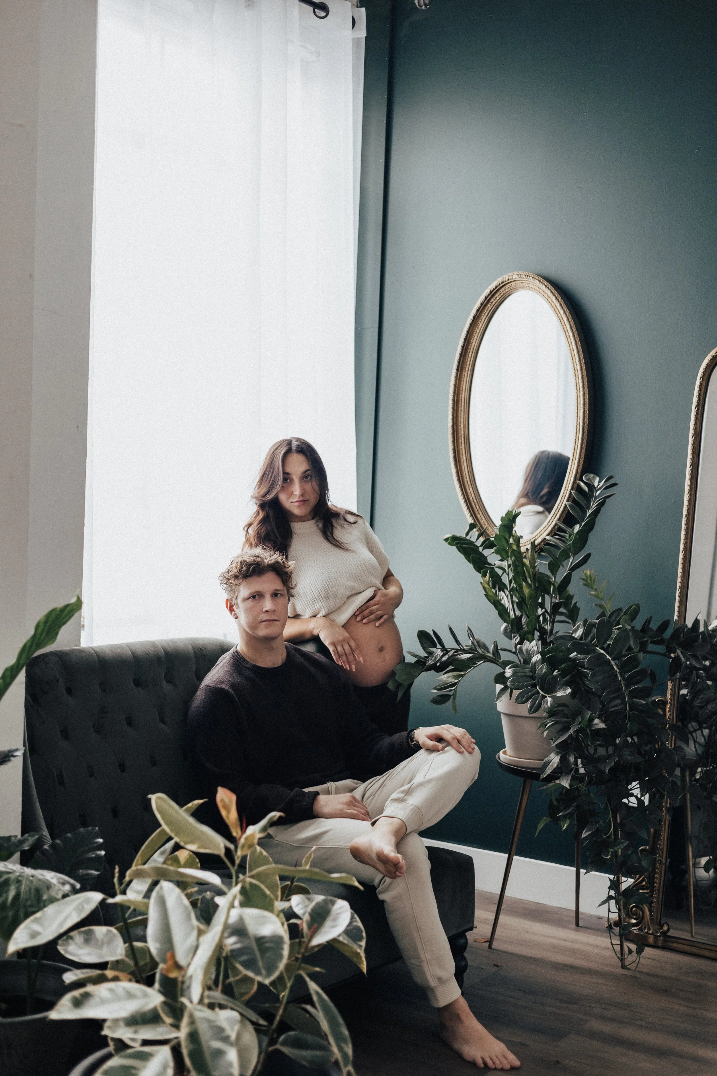 A pregnant woman standing behind a seated man in an indoor setting with green walls, plants, large mirrors, and a window with white curtains. a luxury lifestyle photography photoshoot