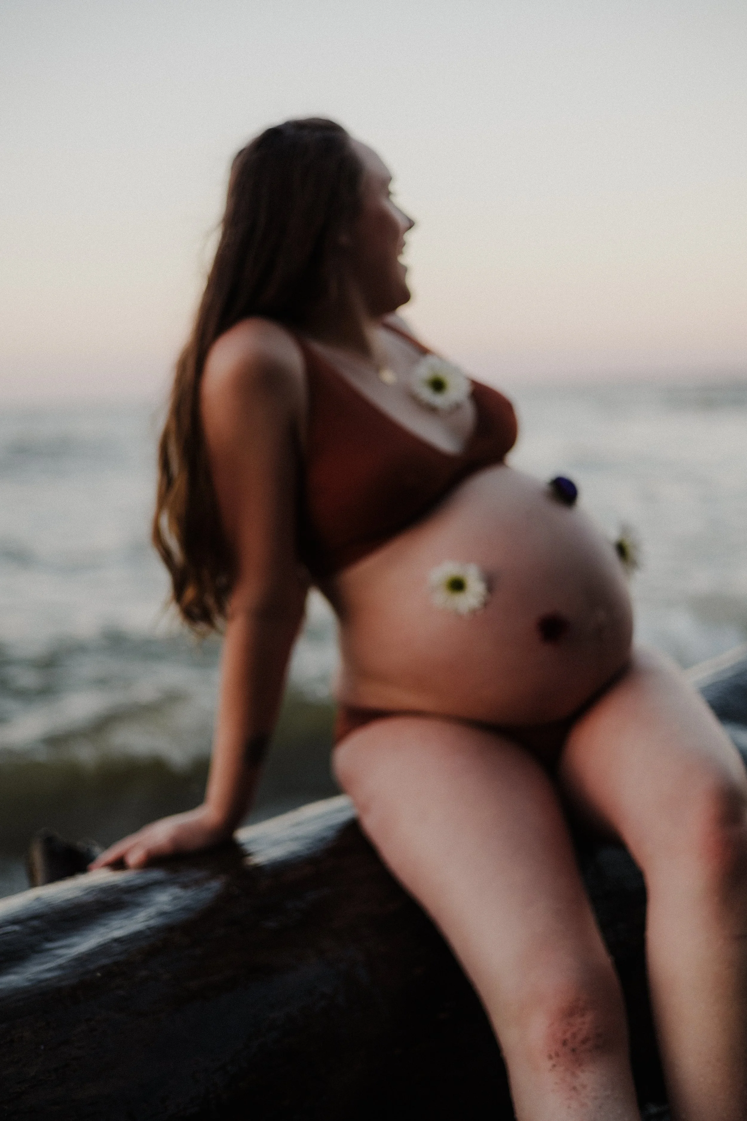 A pregnant woman sitting on a piece of driftwood at the beach during sunset, decorated with flowers on her body.