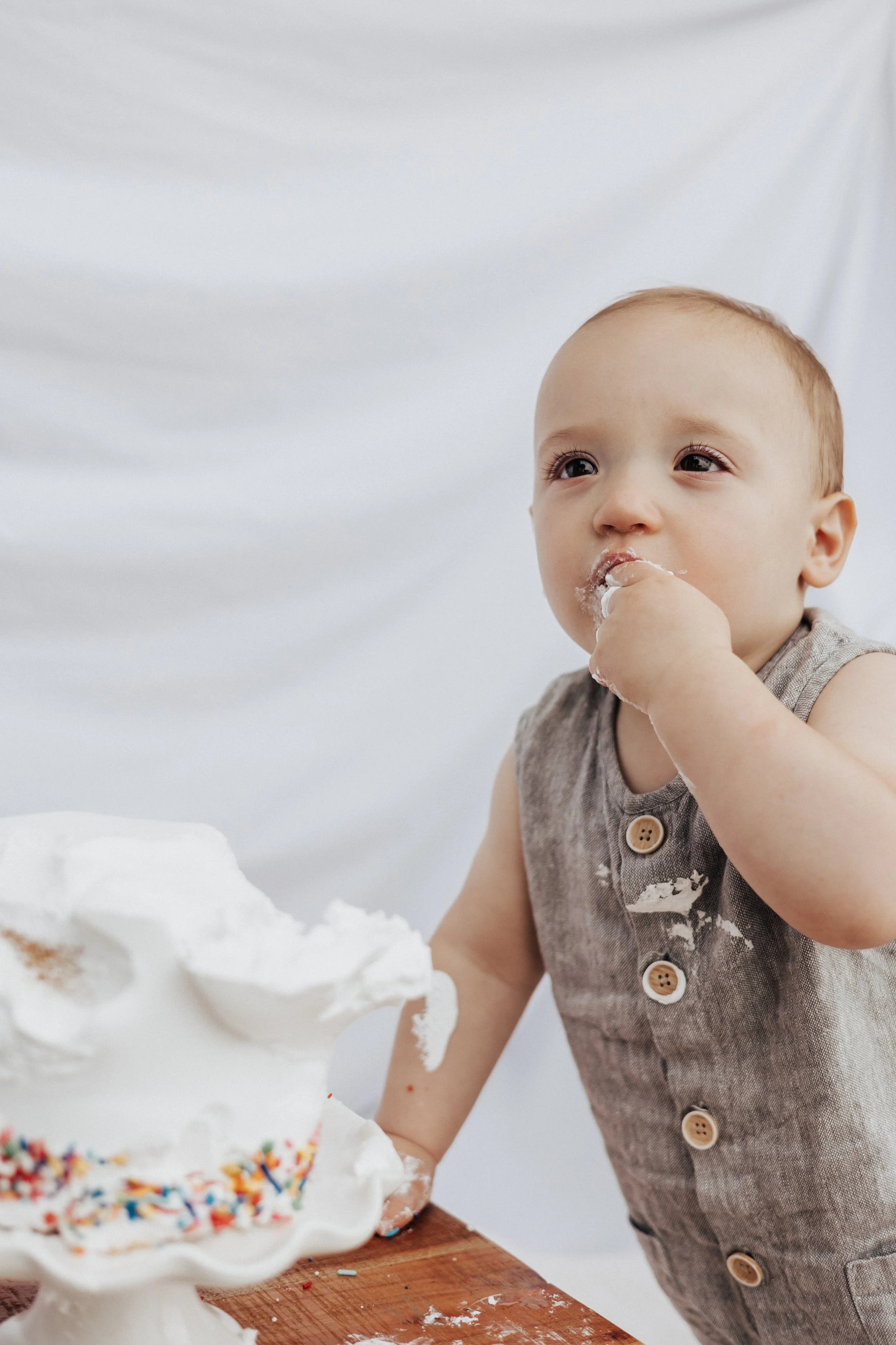 A young child with a piece of cake and rainbow sprinkles on a cake stand, wearing a sleeveless gray button shirt, eating cake with white frosting on face and fingers, standing at a wooden table at a beach in huron ohio