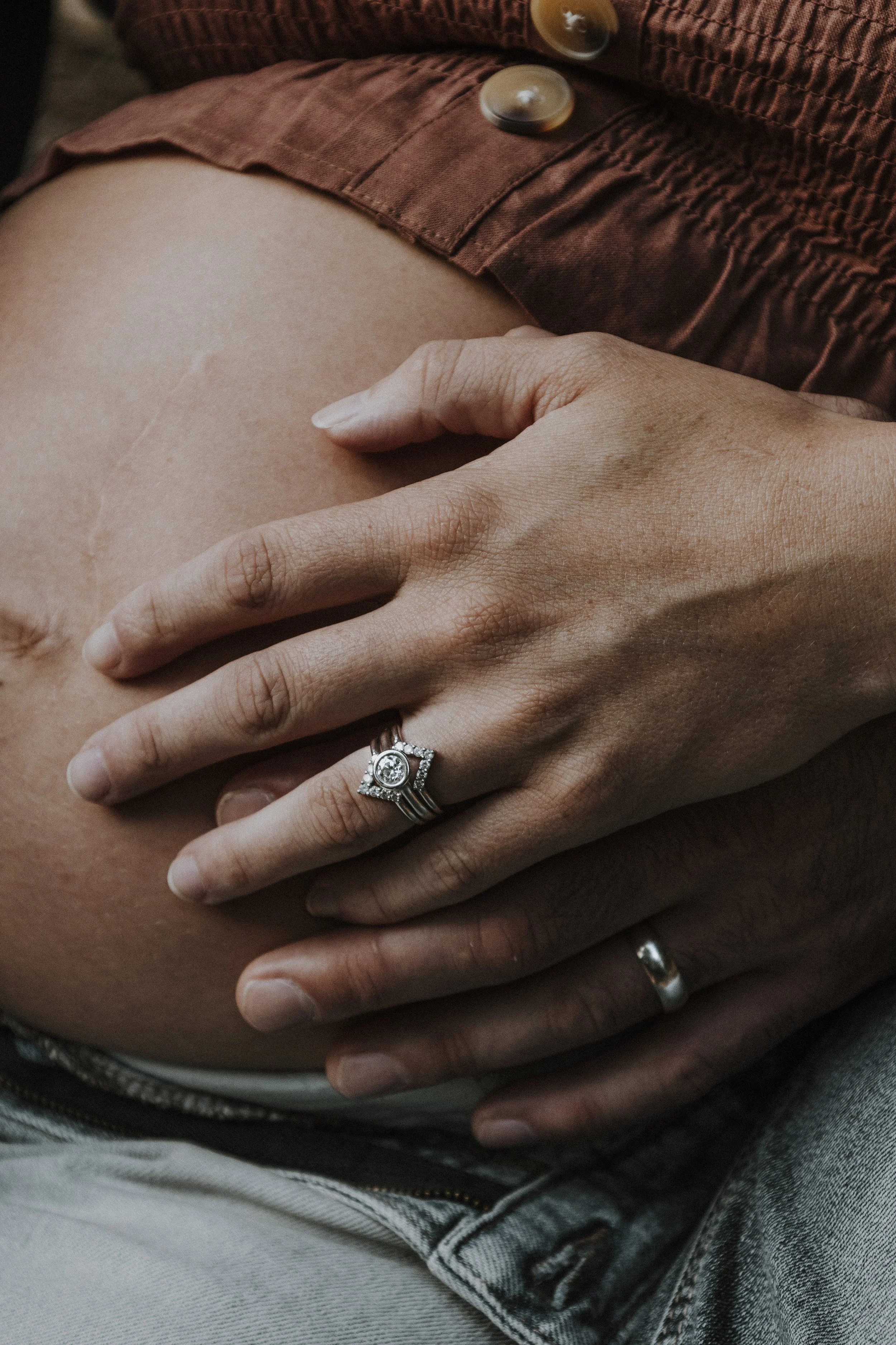 A pregnant person with their hand resting on their belly, showing an engagement ring and wedding band. The person is wearing denim jeans and a reddish-brown top with buttons.