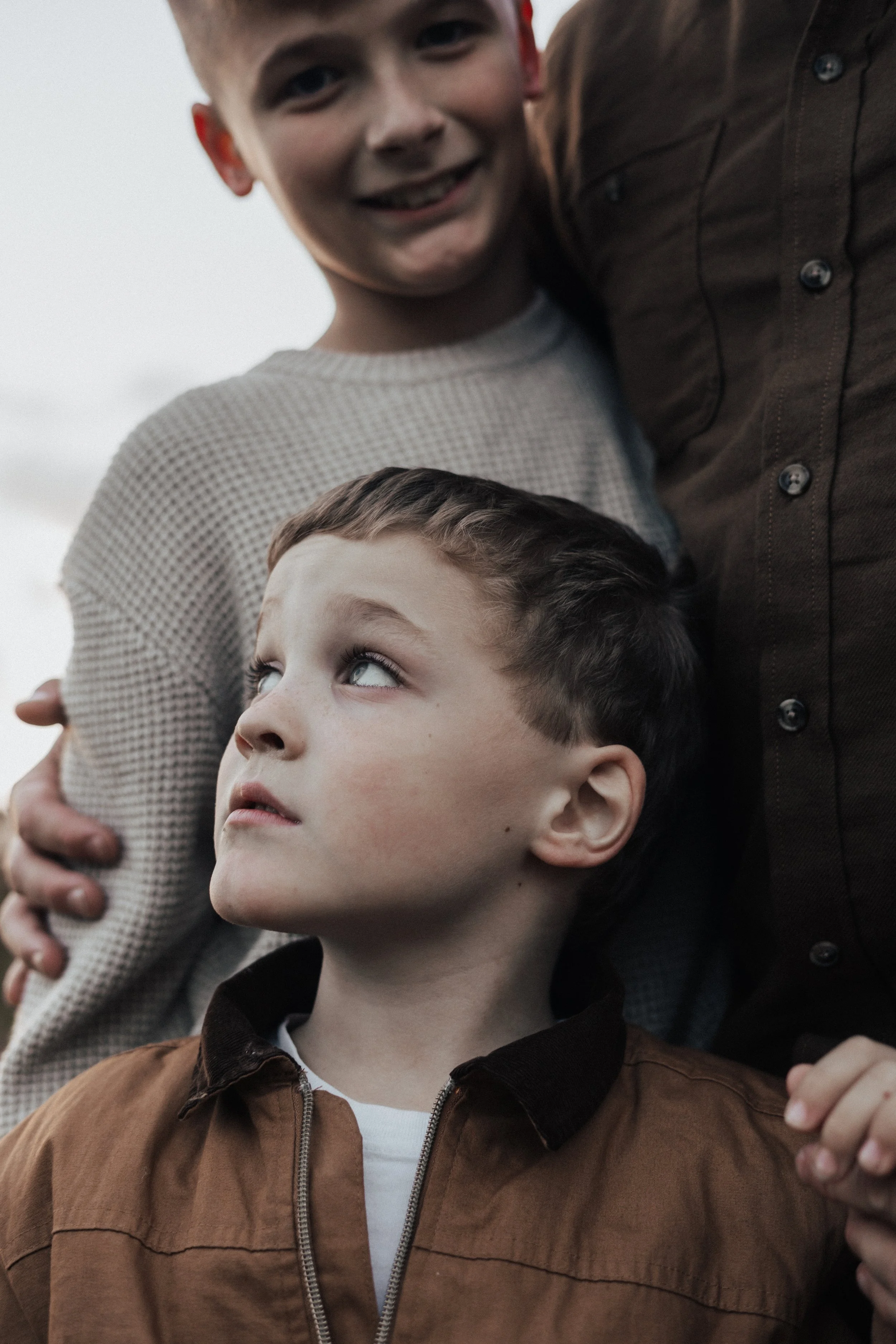 Two boys, one older smiling and one younger looking to the side, with a person partially visible behind them, outdoors.