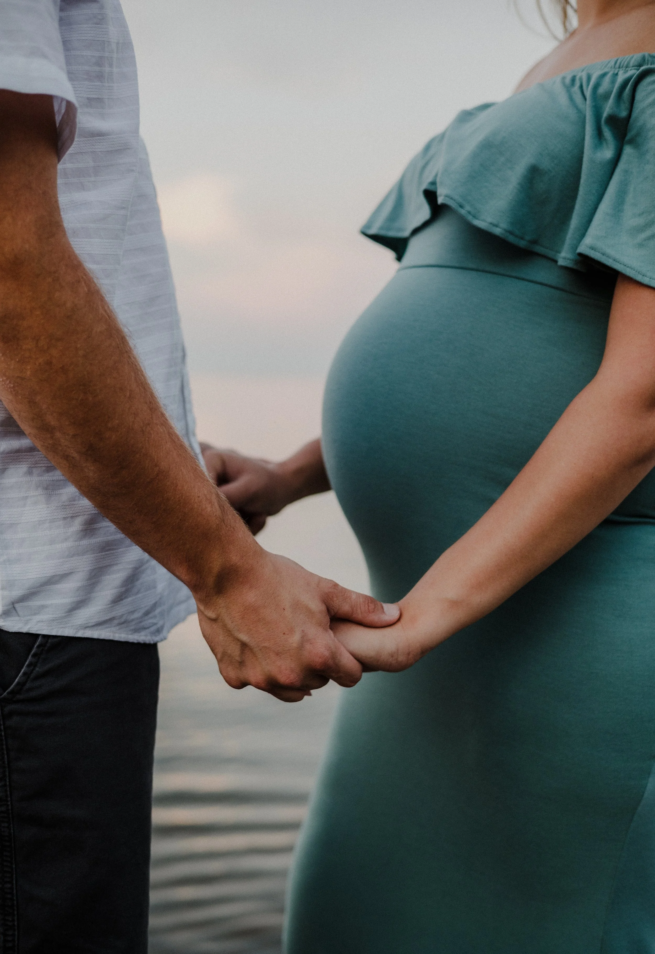 A pregnant woman and a man holding hands near a body of water during sunset.