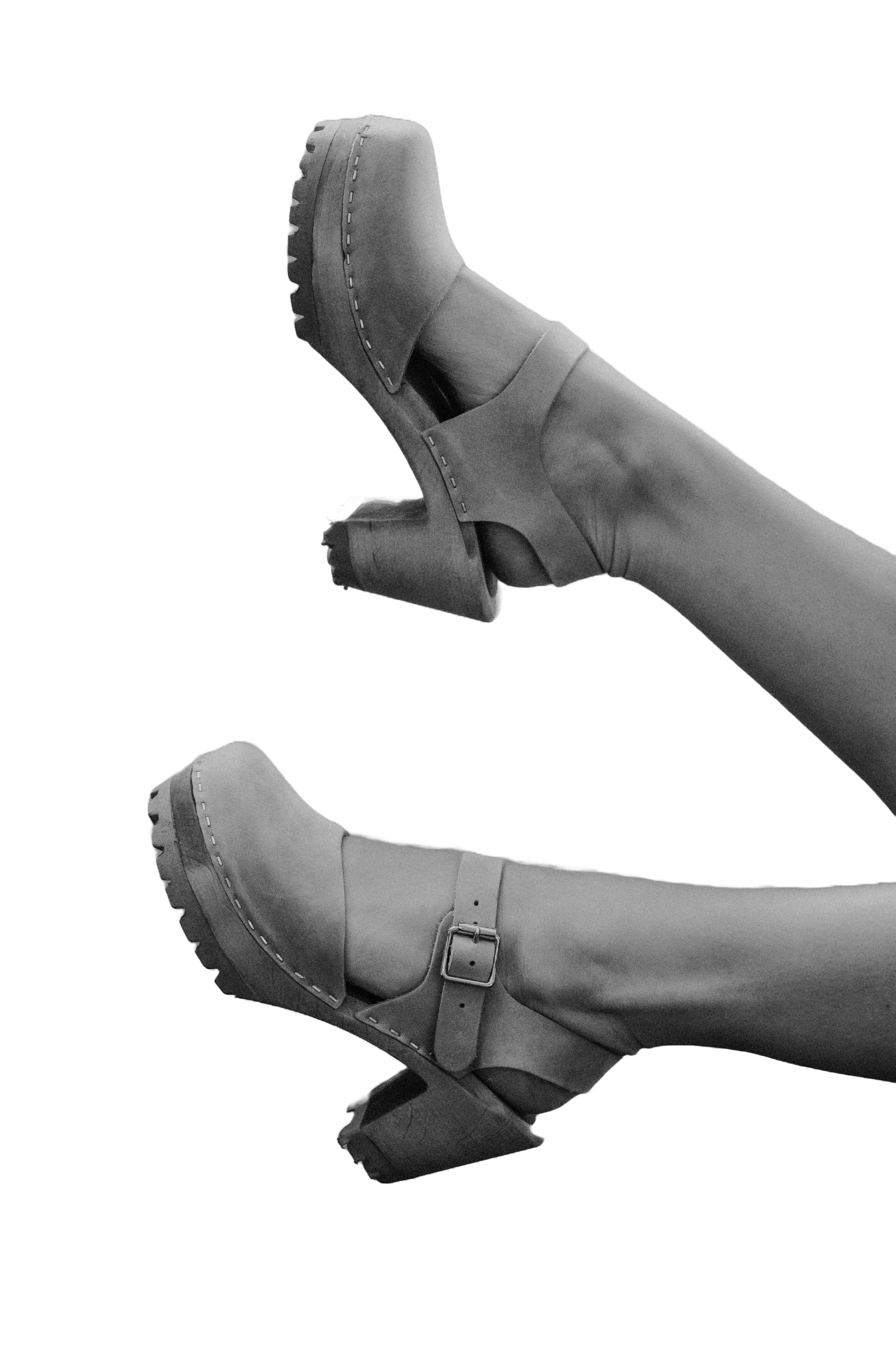 A person wearing high-heeled boots with platform soles, buckles, and straps, shown in black and white for a clog shoe brand