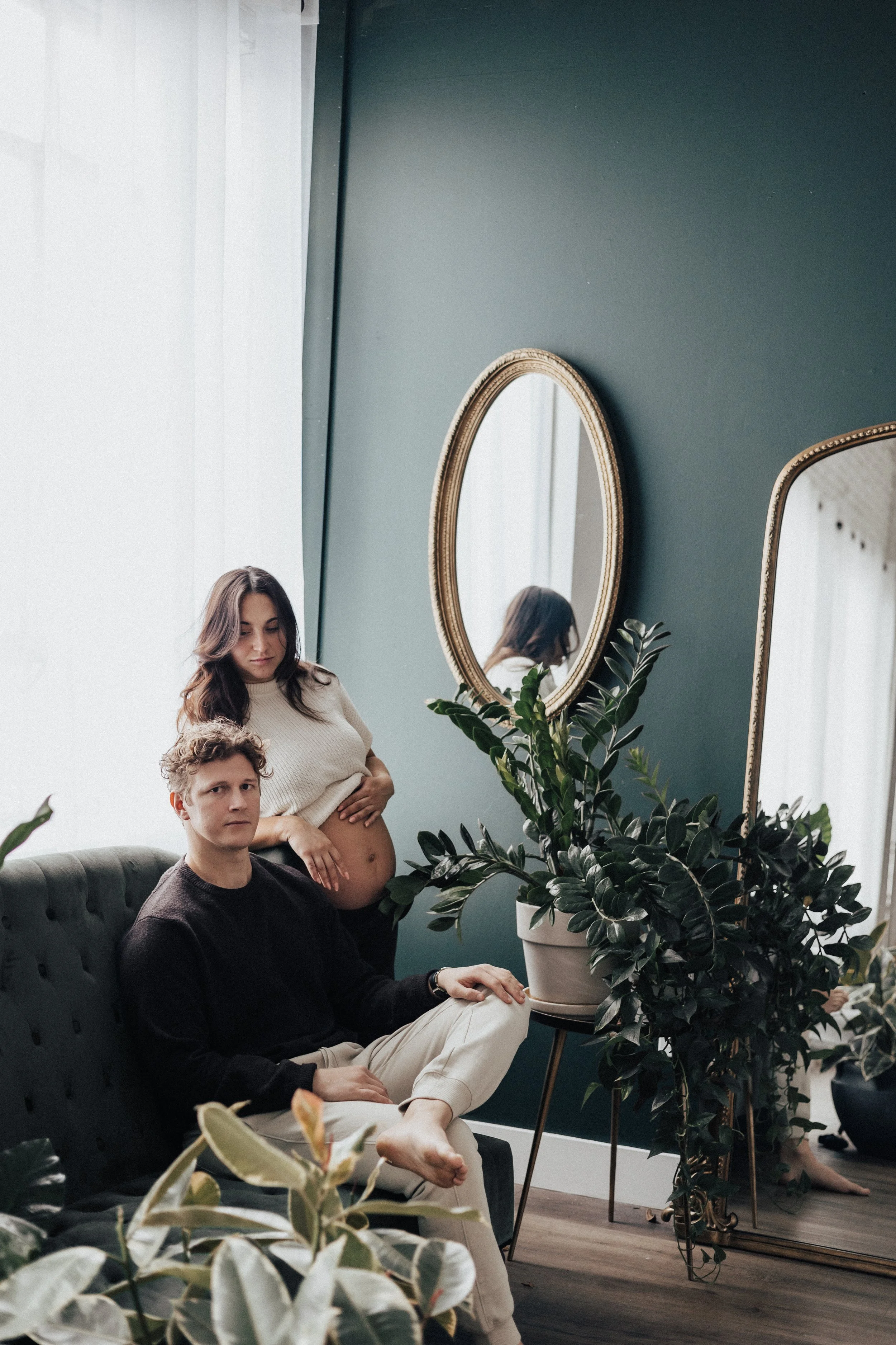 A pregnant woman standing behind a seated man in a room decorated with green walls, large mirrors, and numerous potted plants, including a large plant on a stand.