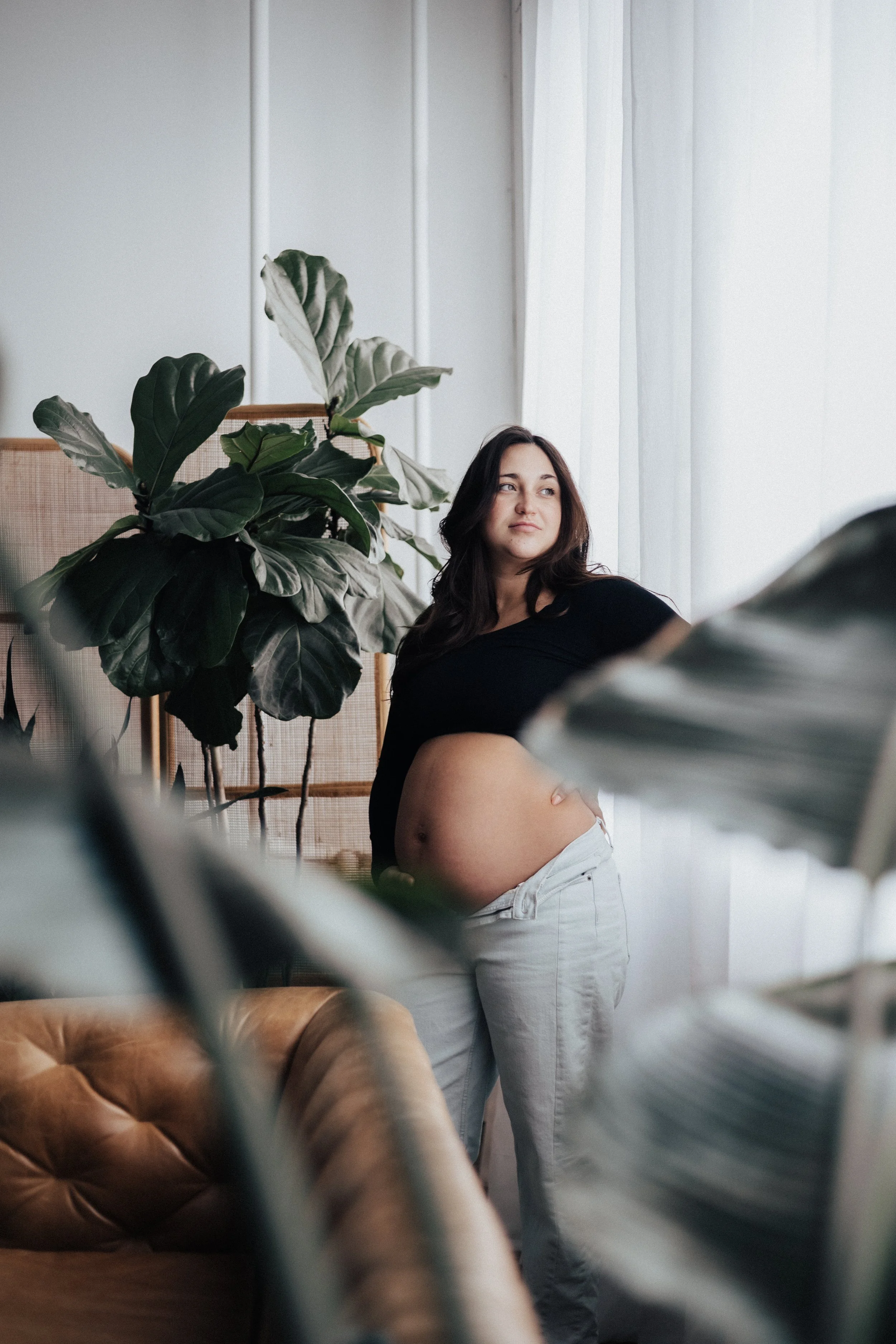 A pregnant woman standing near a window with sheer curtains, wearing a black top and white pants, with a large green plant and a tan leather sofa in the background.