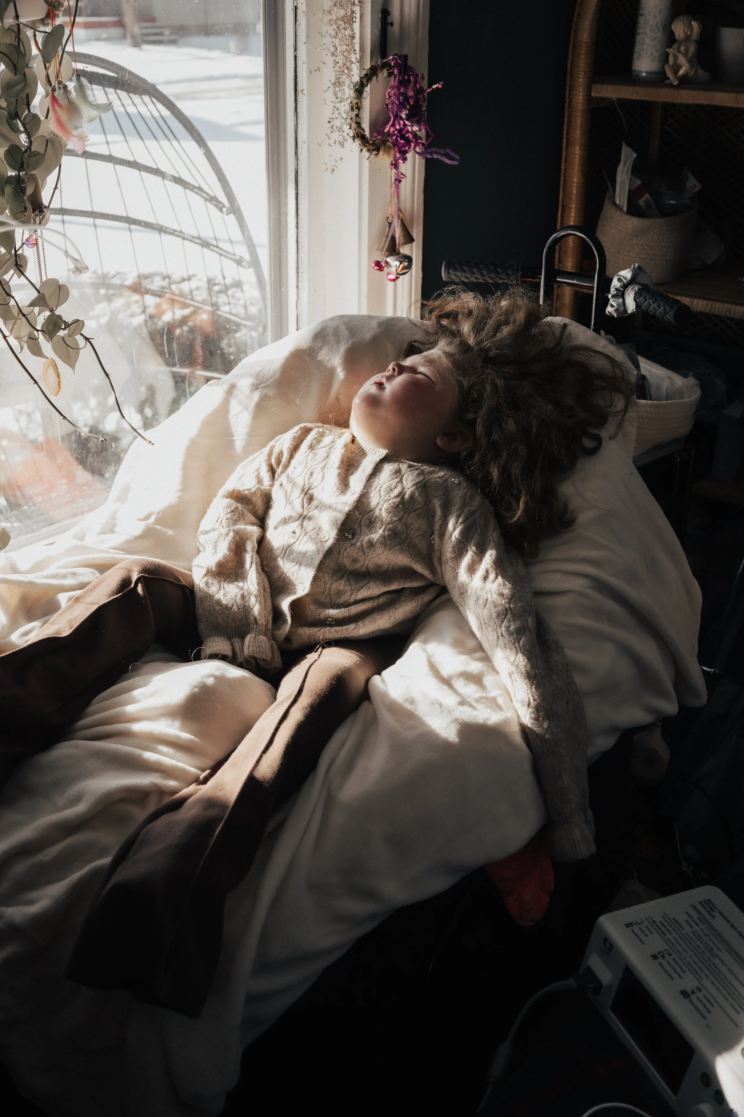 A disabled young child with curly hair sleeping on a bed in front of a window with sunlight streaming in during an in home family photography session in Norwalk ohio