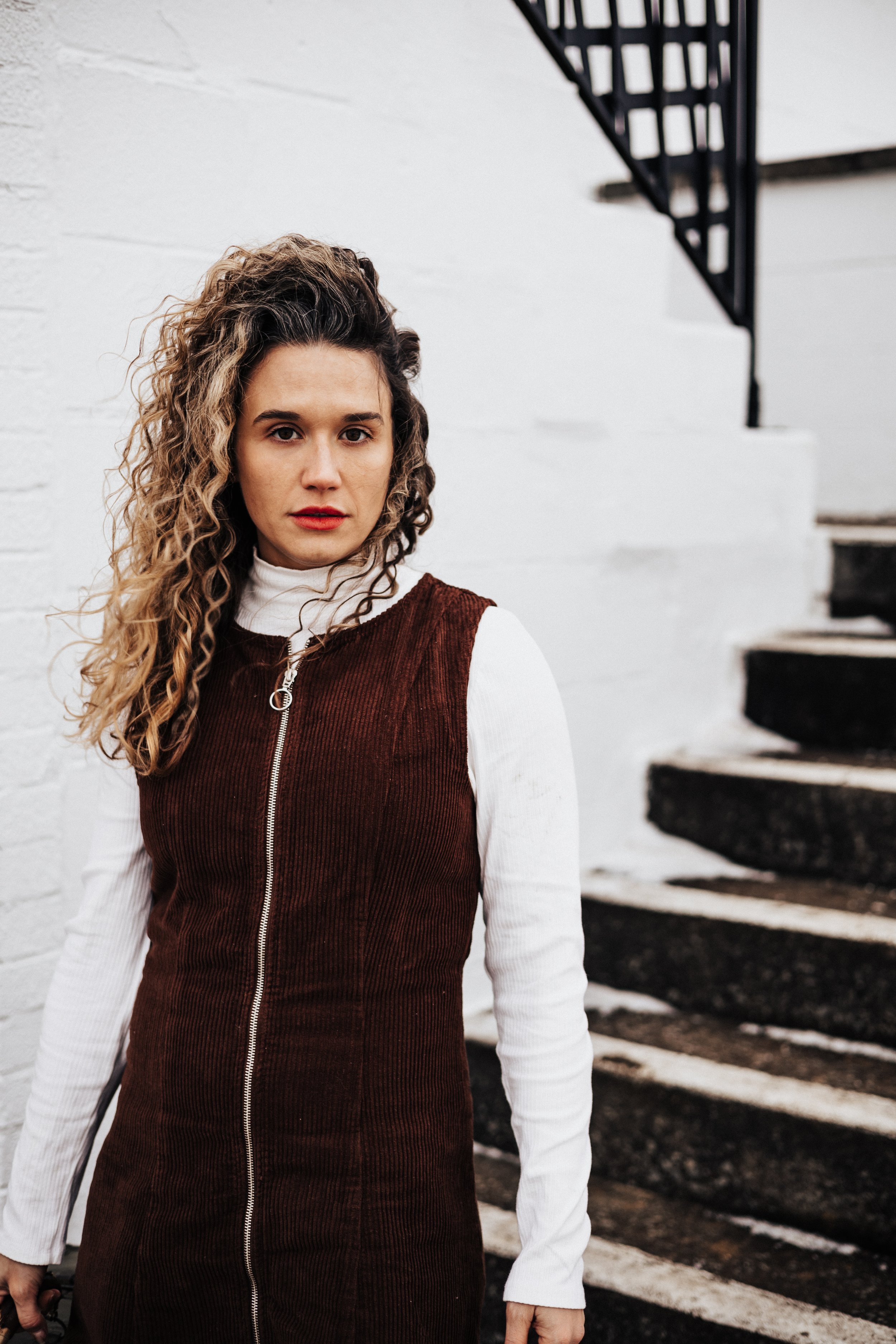 A woman with curly hair standing in front of a white brick wall and black staircase, wearing a white turtleneck and a brown vest with a zipper for a brand at a truckers motel in ohio