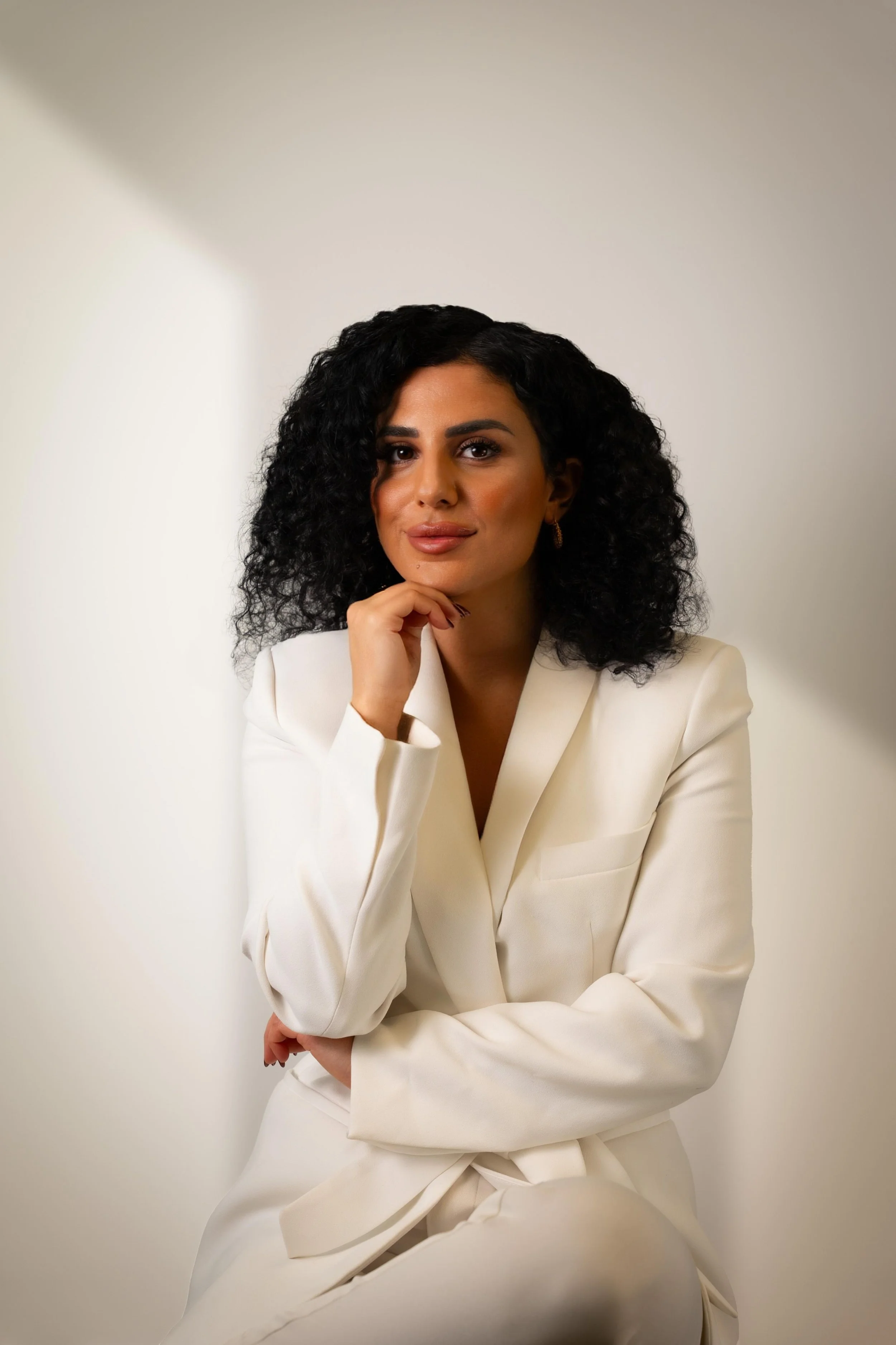 Frau mit lockigem schwarzen Haar in weißem Anzug sitzt vor hellgrauem Hintergrund, schaut in die Kamera, hält chin auf Hand, lächelt leicht.