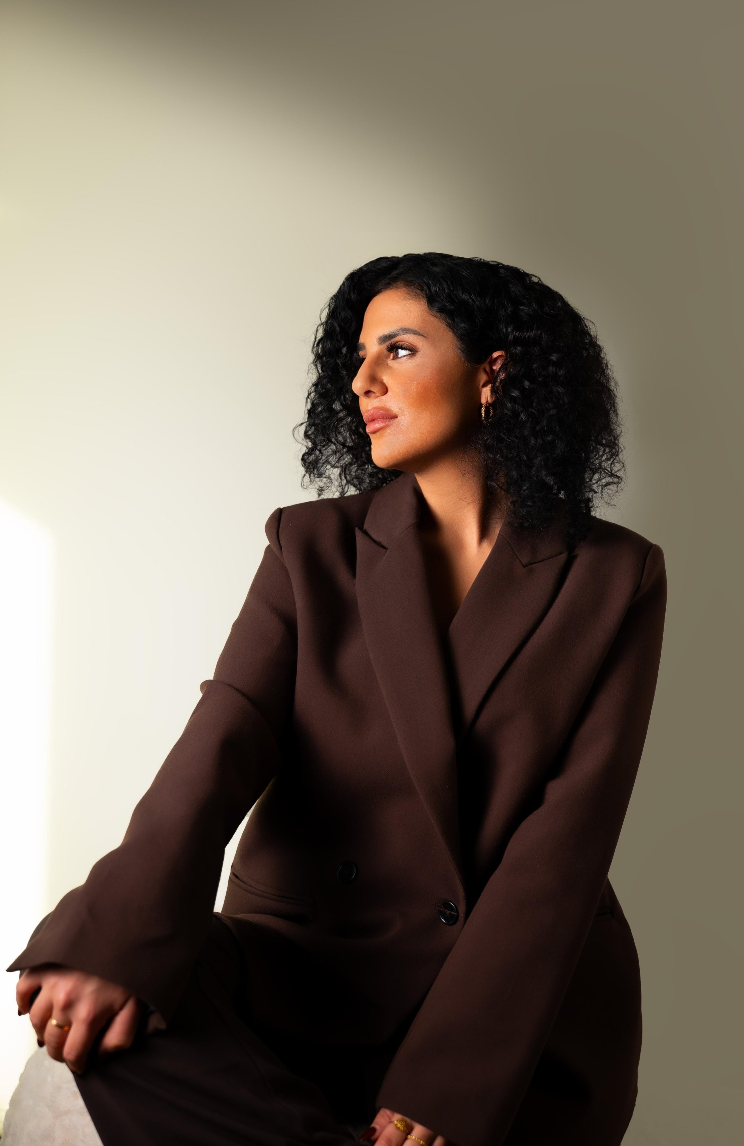 Eine Frau mit lockigem schwarzen Haar trägt einen braunen Blazer und sitzt nachdenklich vor einem neutralen Hintergrund.