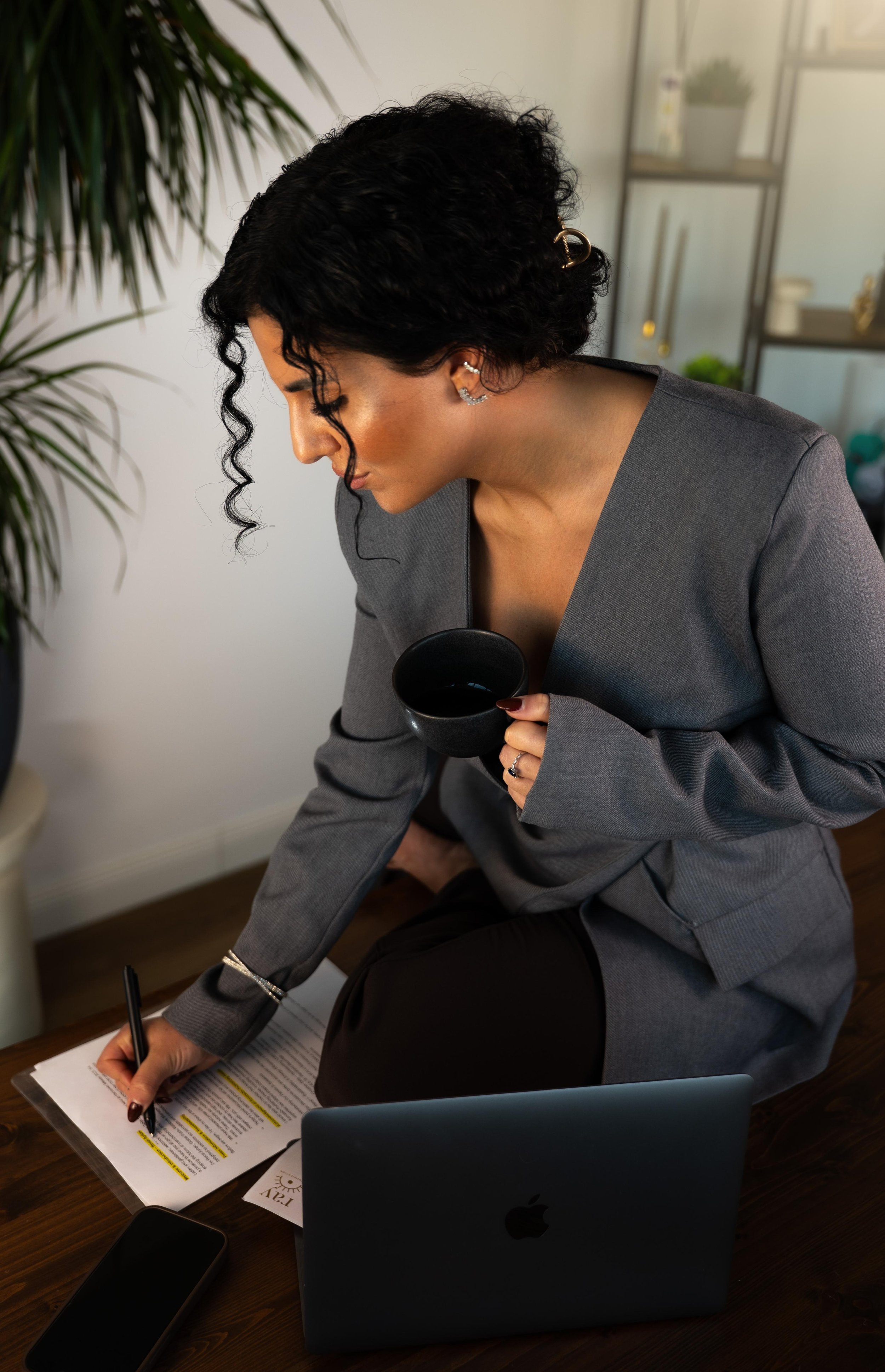 Eine Frau in einem grauen Blazer sitzt auf dem Boden eines Büros, schreibt auf Unterlagen mit einem schwarzen Stift, hält eine schwarze Tasse und arbeitet an einem Laptop.