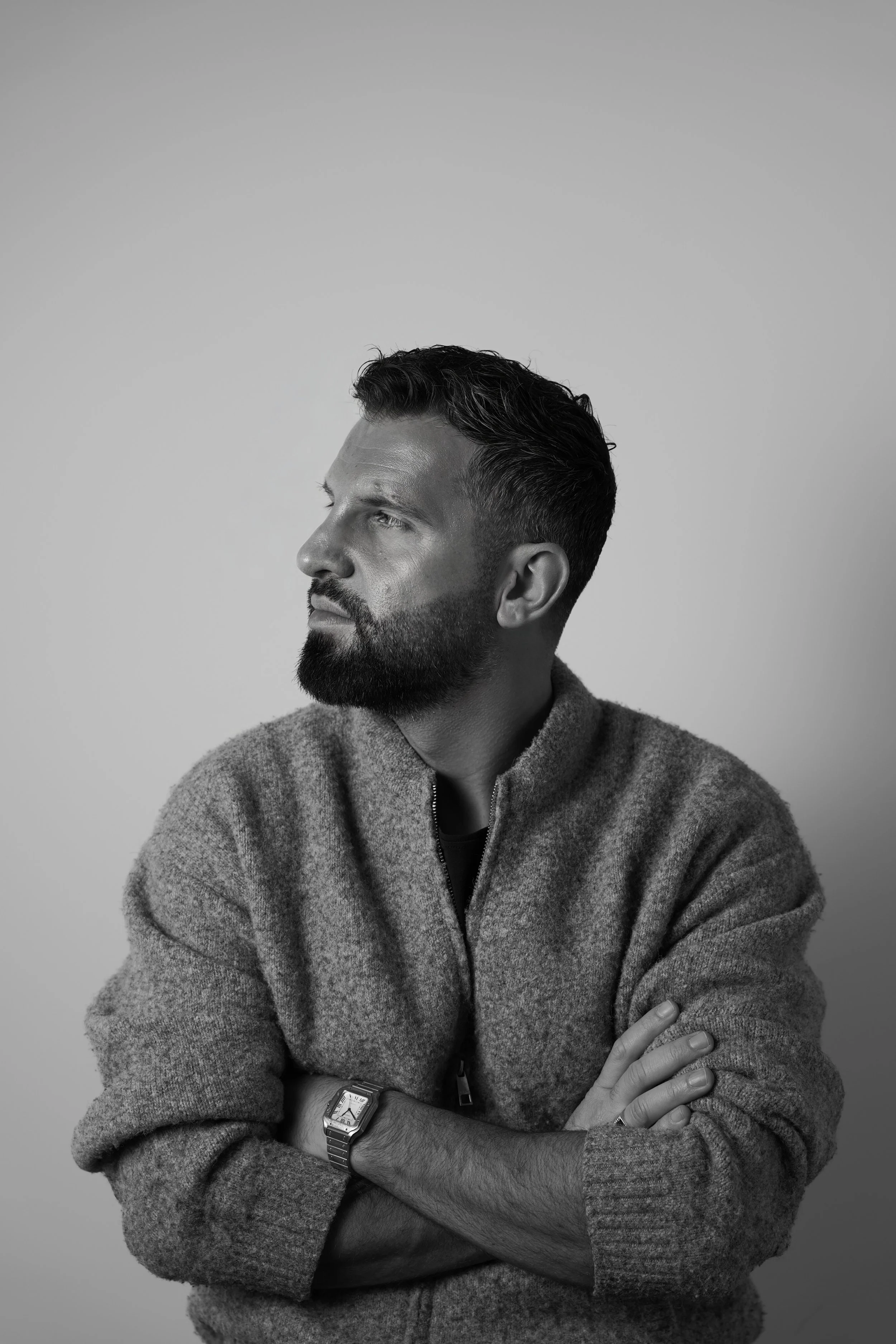 Schwarz-Weiß-Foto eines Mannes mit Bart, der nach links schaut, Arme verschränkt, trägt eine graue Jacke und eine Armbanduhr.