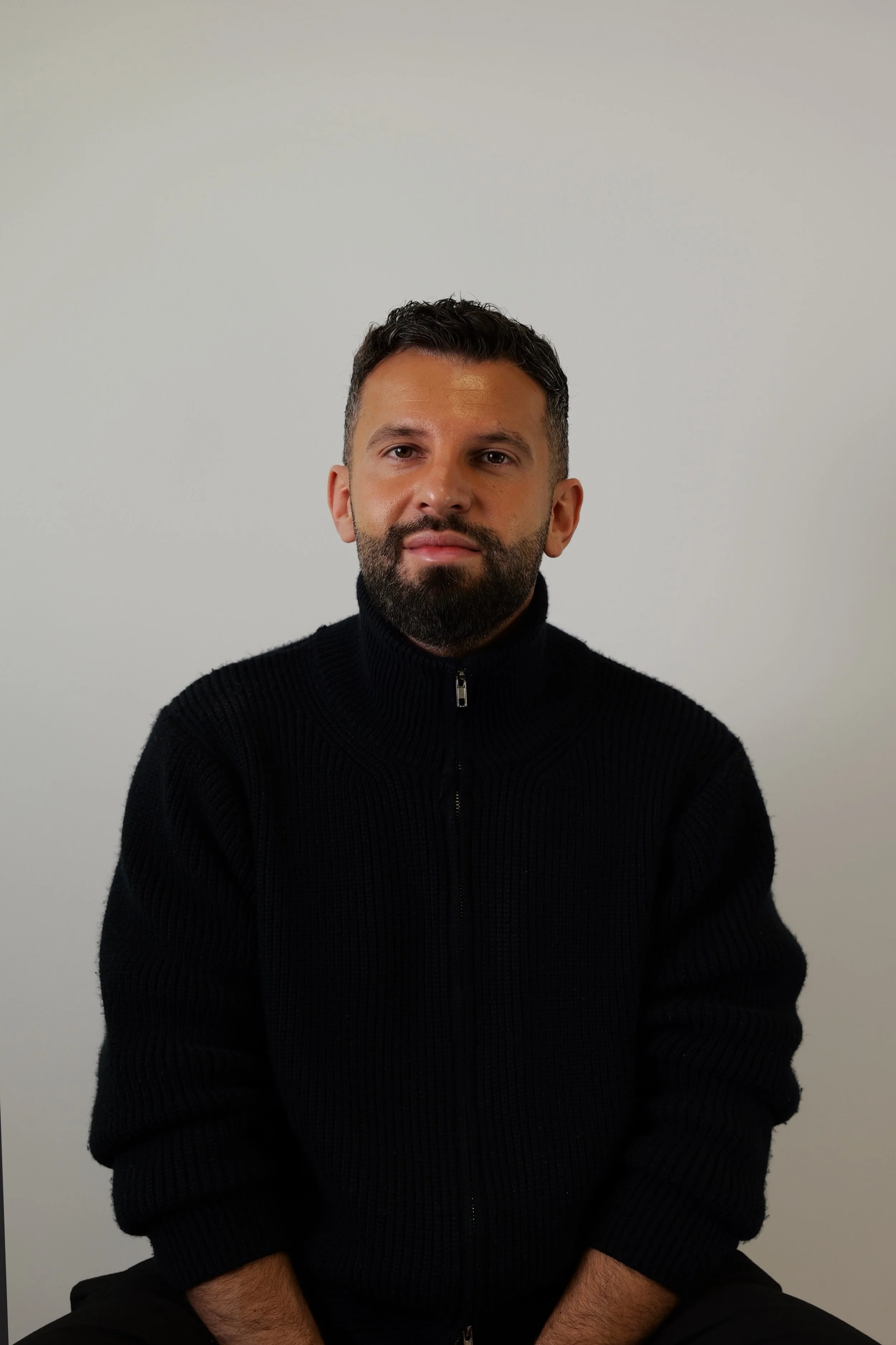 Porträt eines Mannes mit dunklem, kurz geschnittenem Haar, Bart und dunkelrotem Teint, der schwarze Pullover trägt. Er sitzt vor einem weißen Hintergrund.
