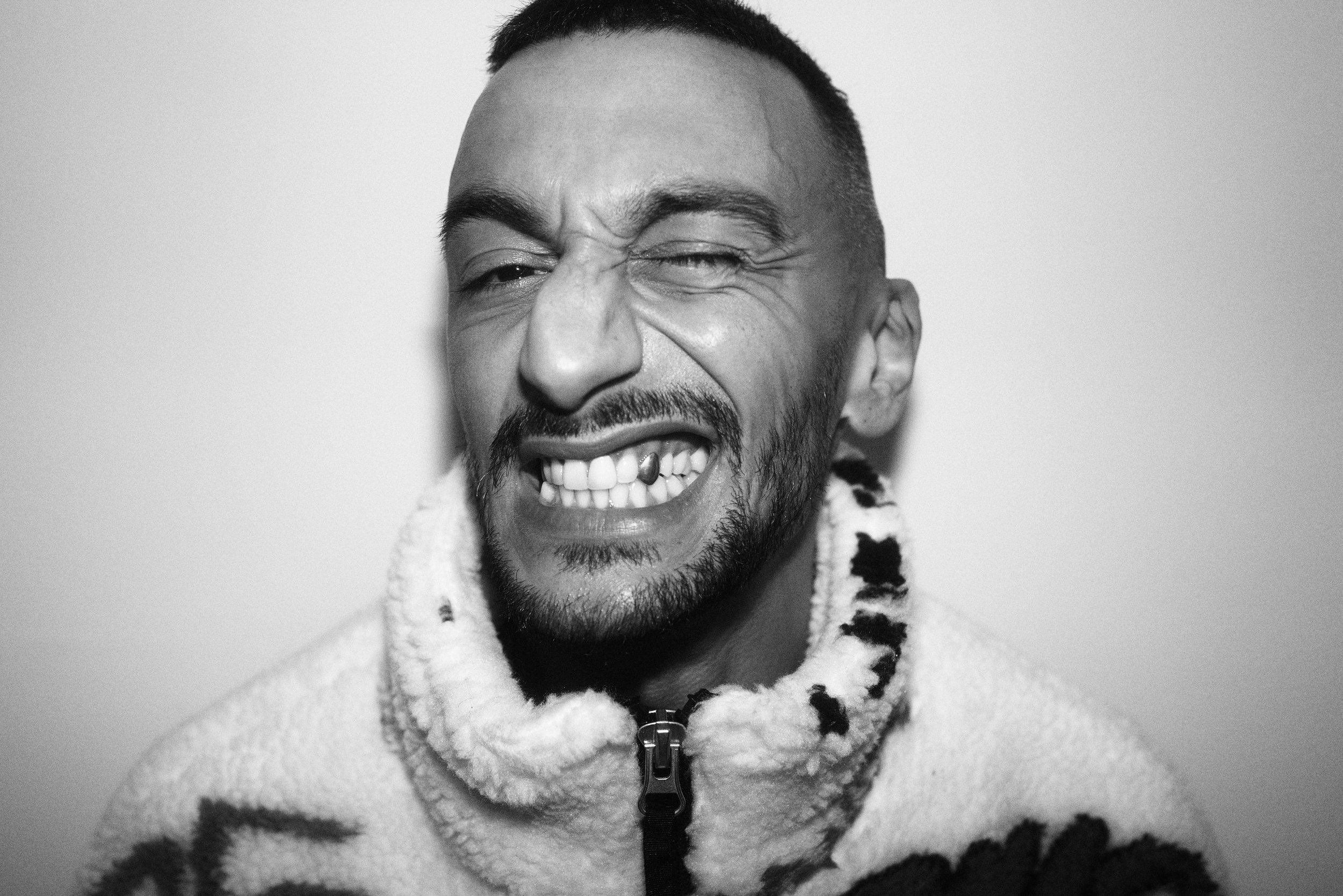 Schwarz-Weiß-Foto eines lachenden Mannes mit zotteligem Pullover, fast komplett zu erkennen, nur ein Auge halb geschlossen.