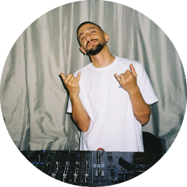 Mann steht vor einem DJ-Setup, trinkt aus einer Flasche und macht eine smilende Geste mit beiden Händen.