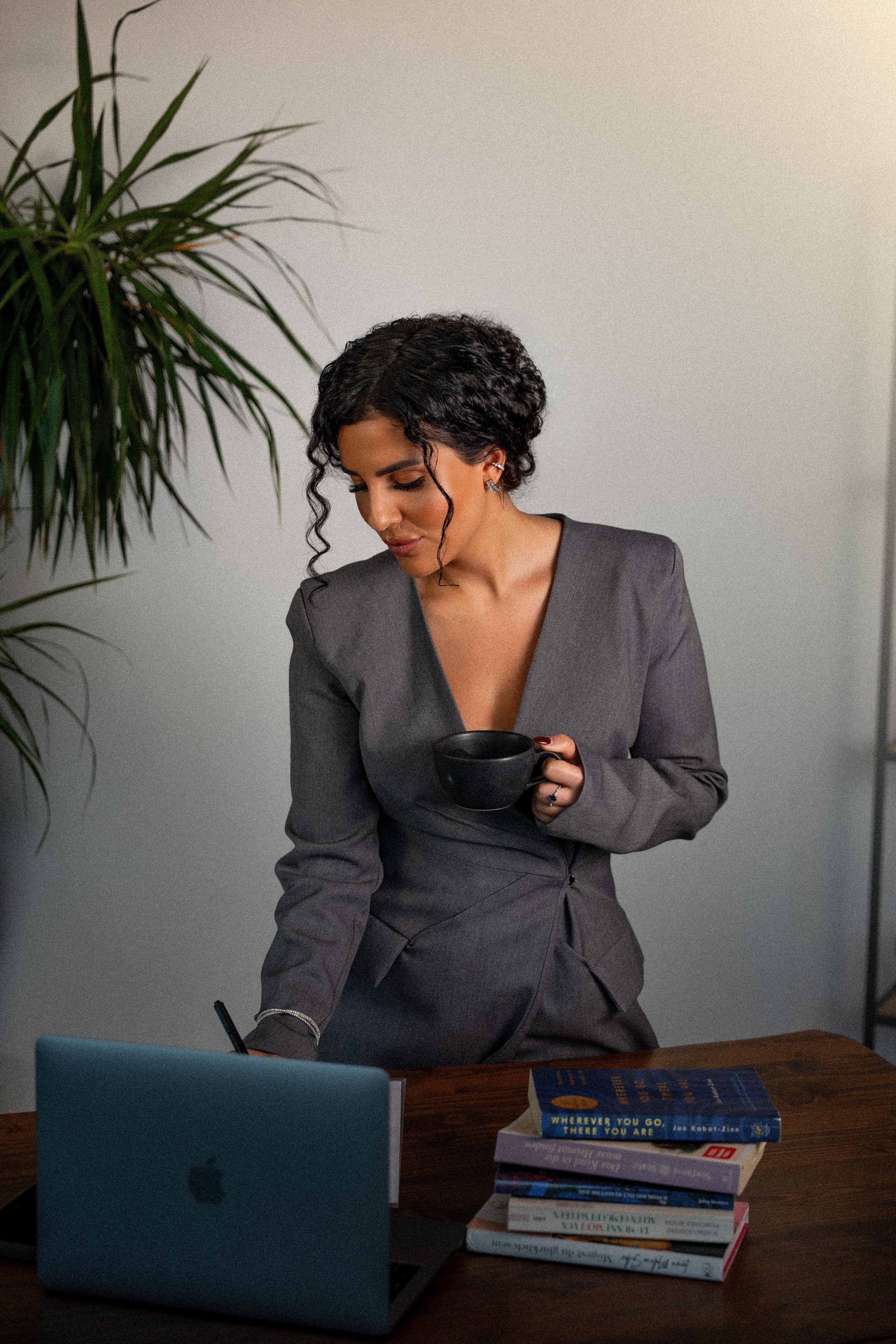 Eine Frau in einem grauen Business-Anzug steht an einem Schreibtisch mit einem Laptop, mehreren Büchern und einer Tasse in der Hand, während sie in die Nähe des Bildschirms schaut.