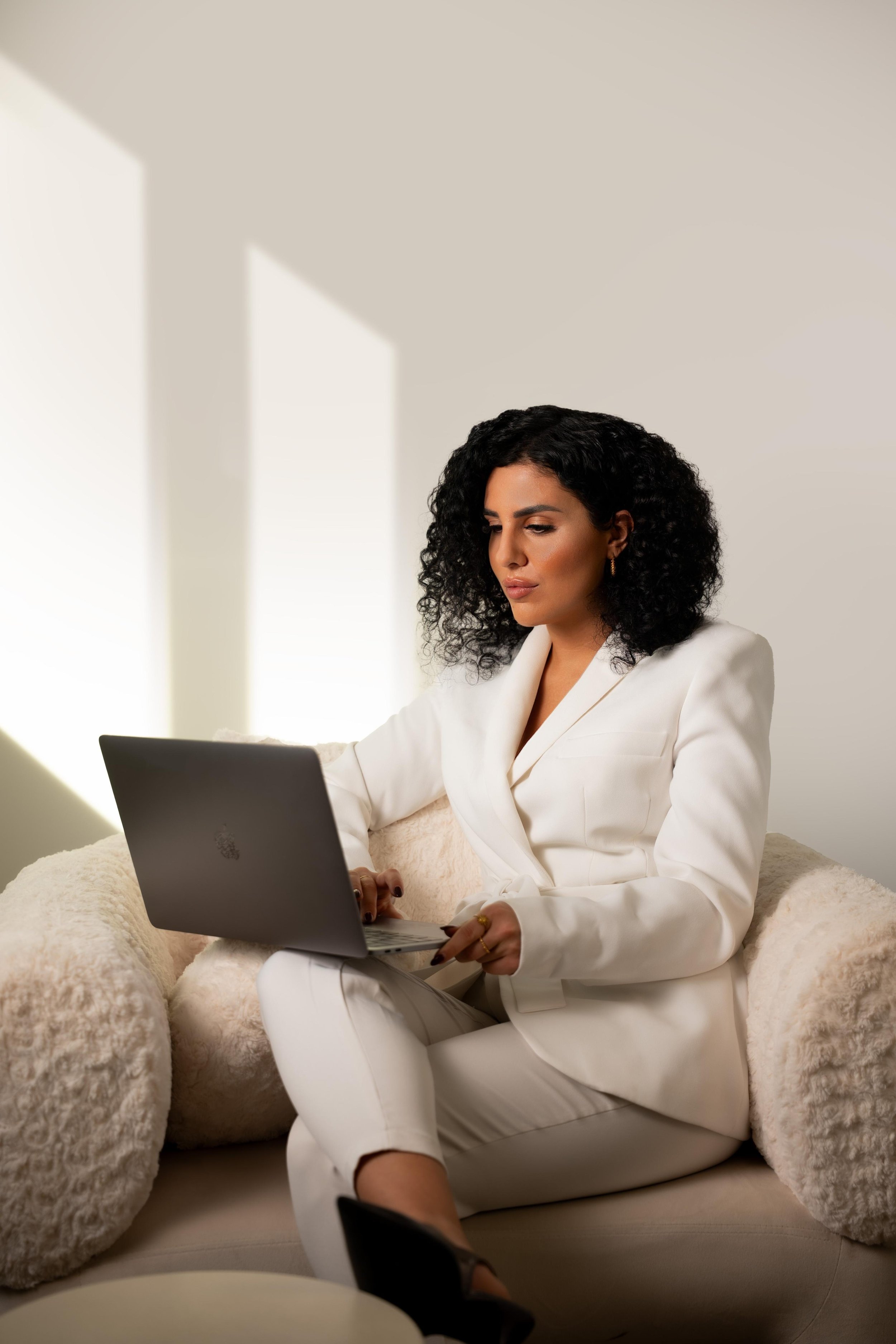 Frau in weißem Anzug arbeitet mit einem Laptop auf einem gemütlichen Sofa, im Hintergrund Sonnenlicht durch Fenster
