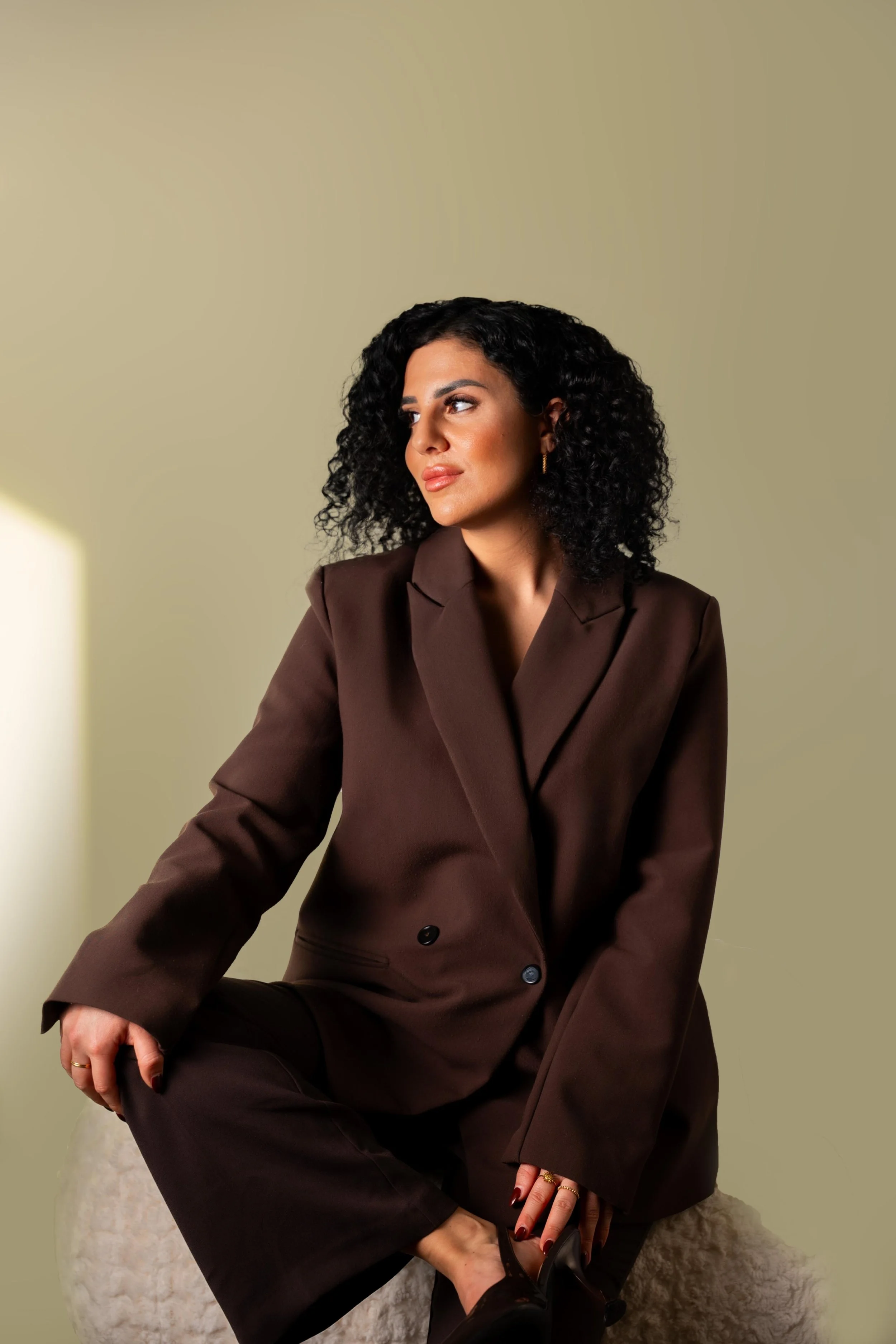 Frau in braunem Anzug sitzt auf einem weißen, flauschigen Hocker vor einem gelben Hintergrund.