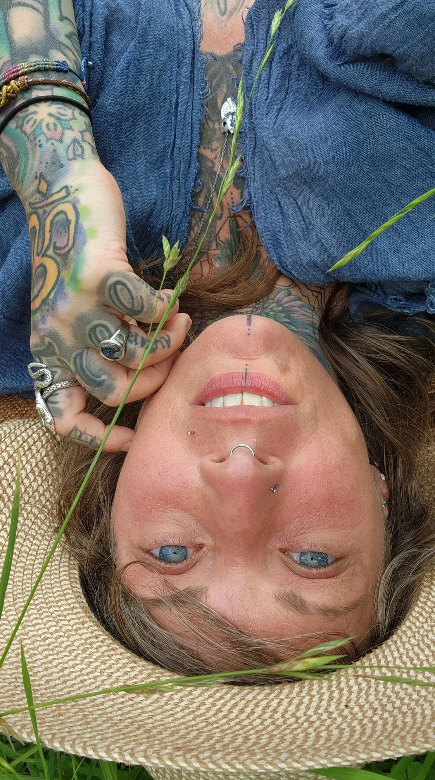 A woman with blue eyes and tattoos lying on her back on grass, wearing a straw hat and a denim shirt. She has piercings on her nose, lips, and ears, and is holding a sprig of grass. Her face has a smile and she is surrounded by green grass.