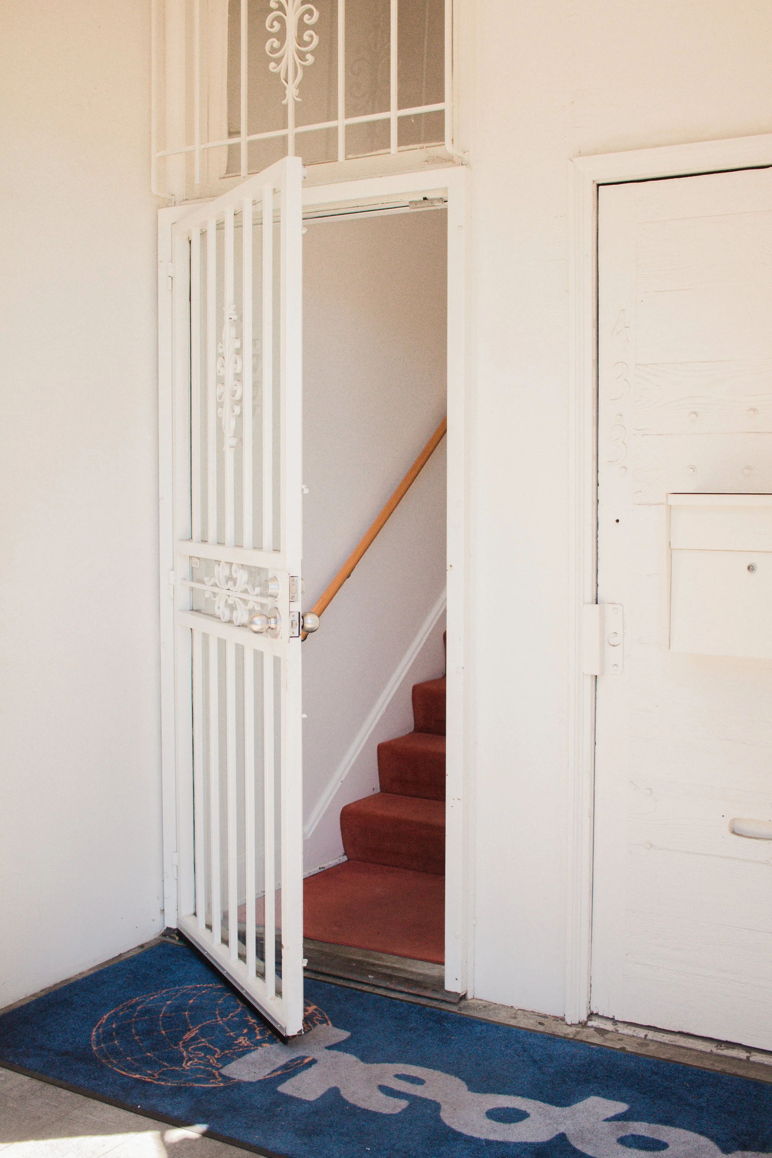 A white metal security door open to reveal a staircase with brown carpet, a beige wall, and a wooden handrail visible. There is a blue floor mat with a circular pattern in front of the door.