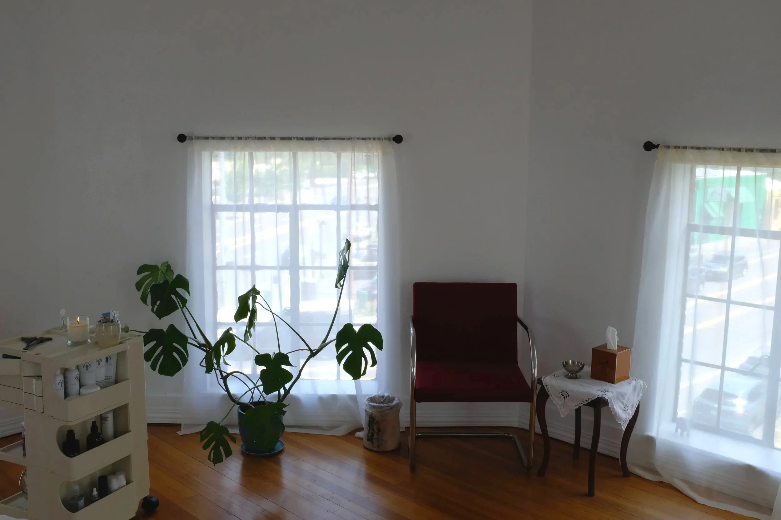 A cozy corner of a room with large windows covered by sheer white curtains. There is a dark-colored armchair, a small table with a lace tablecloth, a wooden tissue box, a candle, and a decorative bowl. A tall houseplant with large, lobed leaves is near the window. The room has hardwood flooring and a bright, airy atmosphere.