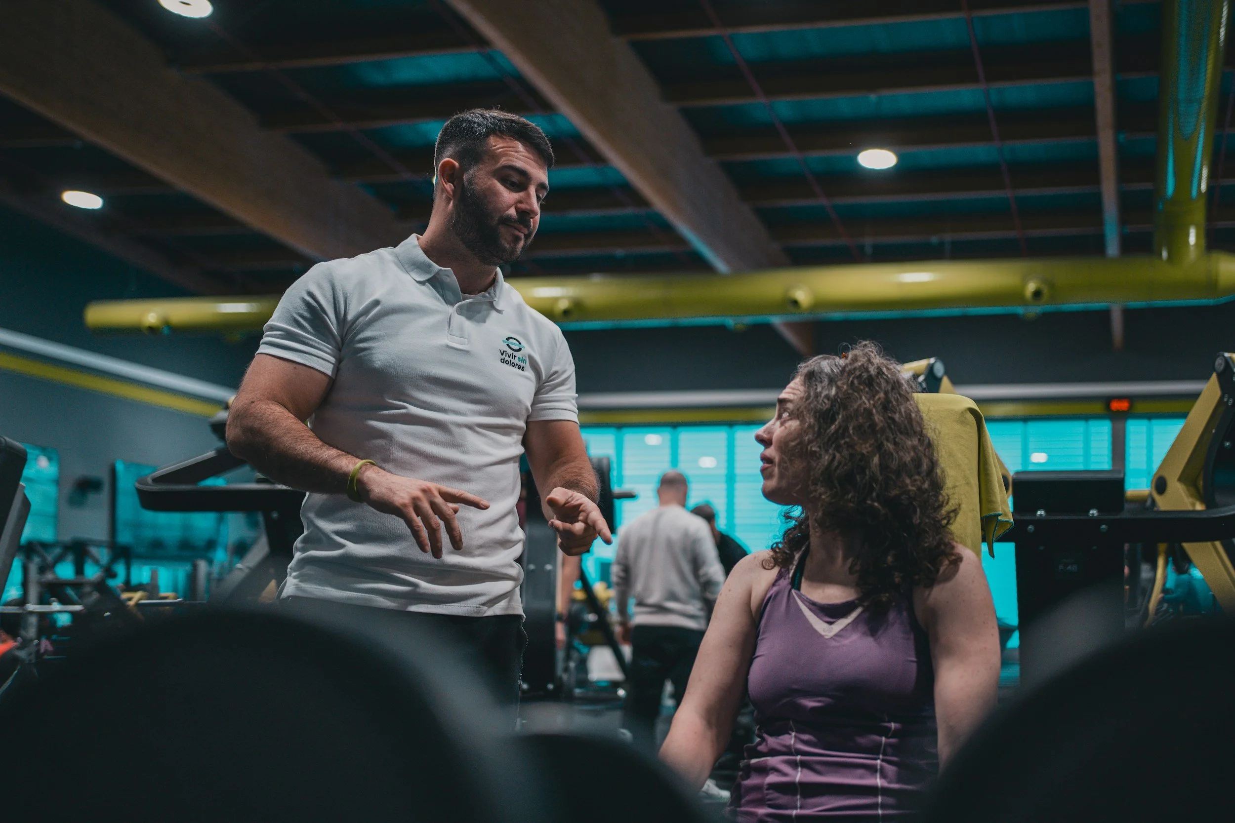 A male personal trainer with dark hair and beard, wearing a white polo shirt with a logo, demonstrating an exercise to a seated woman with curly hair in a gym. The gym has yellow and teal equipment.