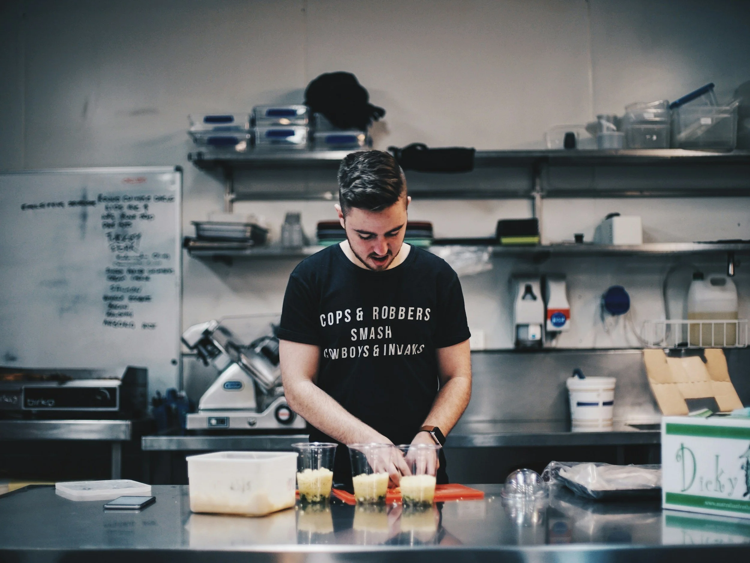 A young man working in a kitchen or bakery, wearing a black t-shirt with white text. He is standing at a workspace with various food items and containers on the counter, with shelves and kitchen equipment in the background.