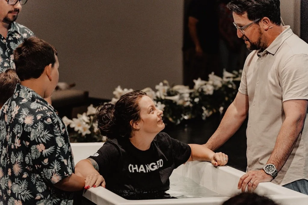 Woman being baptized in a baptismal pool while a man supports her, with children watching closely.