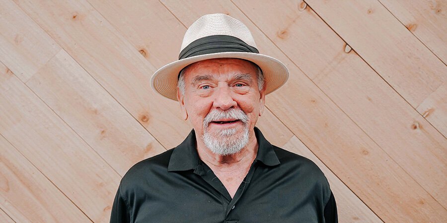 Smiling elderly man with a white beard and mustache, wearing a white straw hat with a black band and a black shirt, standing against a light wooden wall.