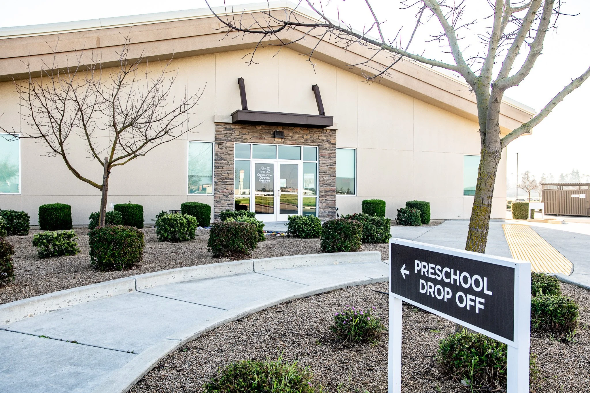 Exterior view of a preschool drop-off area with a black sign indicating 'Preschool Drop Off' pointing left. The building has a beige facade with stone accents around the entrance, large glass doors, and windows. There are leafless trees, shrubs, and a sidewalk with a curved curb.