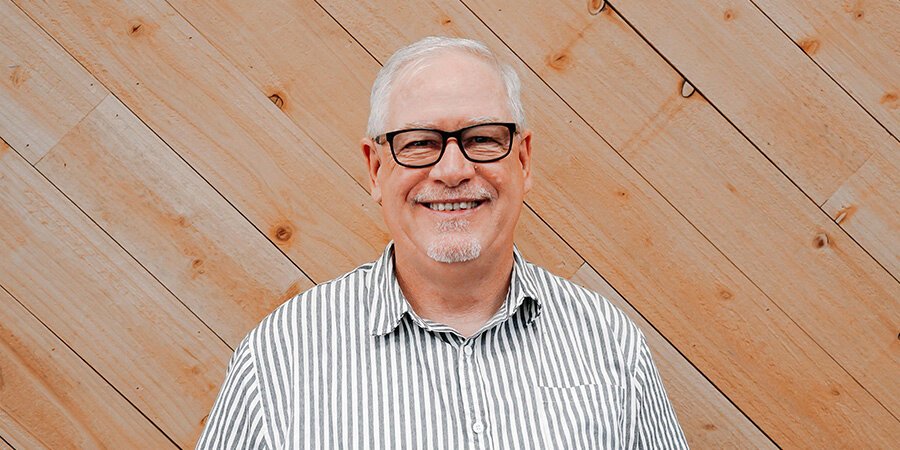 A smiling man with glasses and white hair, wearing a striped shirt, standing against a wooden plank wall.