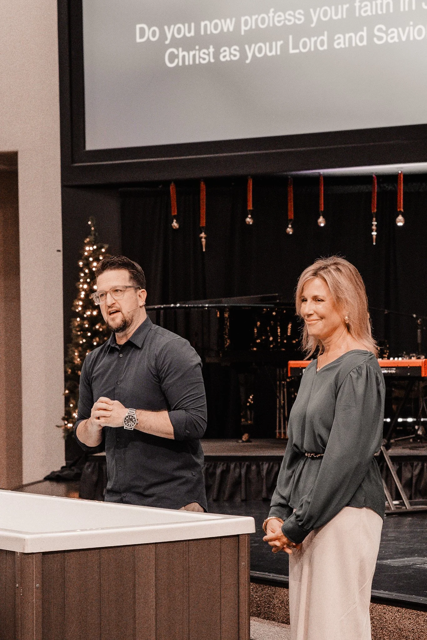 A man and a woman are standing in front of a stage with a black piano, decorated with a Christmas tree with lights in the background. The man is speaking, wearing glasses and a dark shirt, while the woman is smiling, wearing a green blouse and beige pants.