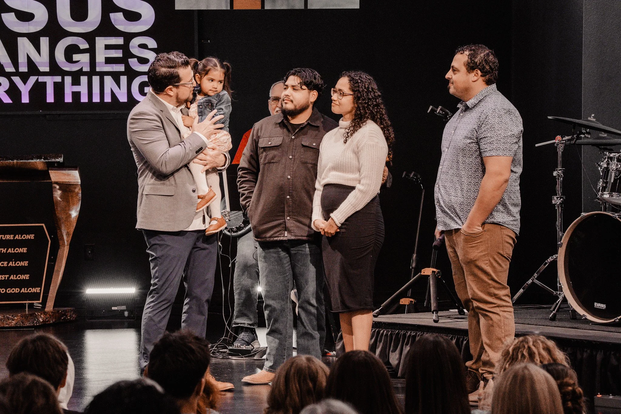 A group of people standing on a stage during a church service or event. A man in a gray suit is holding a young girl and speaking, while others listen. The stage has musical instruments in the background.