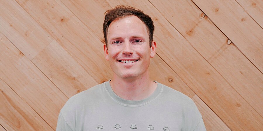 A smiling man with short brown hair wearing a light gray T-shirt, standing in front of a wooden wall with diagonal planks.