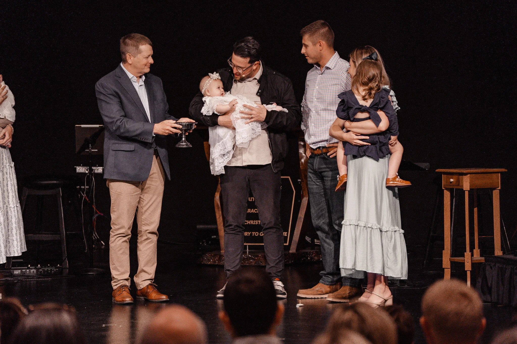 A man holding a baby girl in a white dress with lace, amid a group of adults on a stage during a ceremony or service, against a dark background.