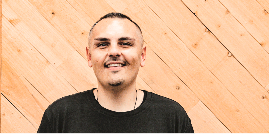 Smiling man with short hair and a goatee, wearing a black T-shirt, standing against a wooden wall background.