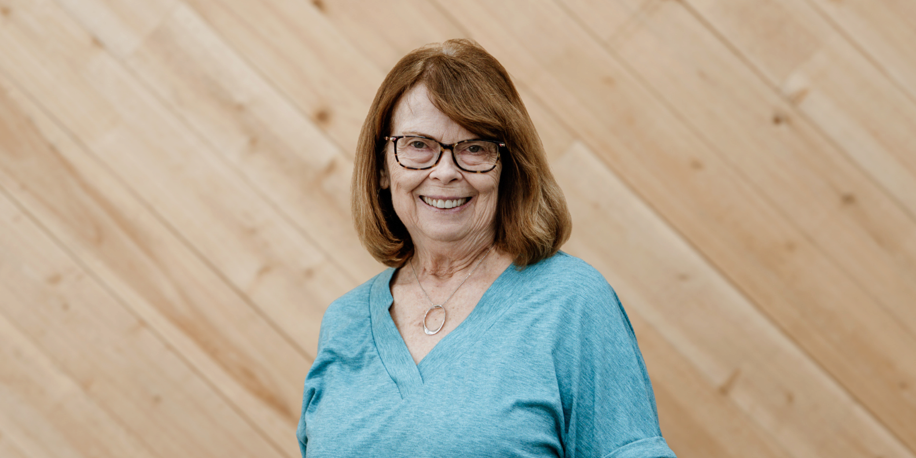 An older woman with glasses and shoulder-length auburn hair smiling at the camera, wearing a light blue V-neck shirt and a silver necklace, standing against a light wooden wall background.