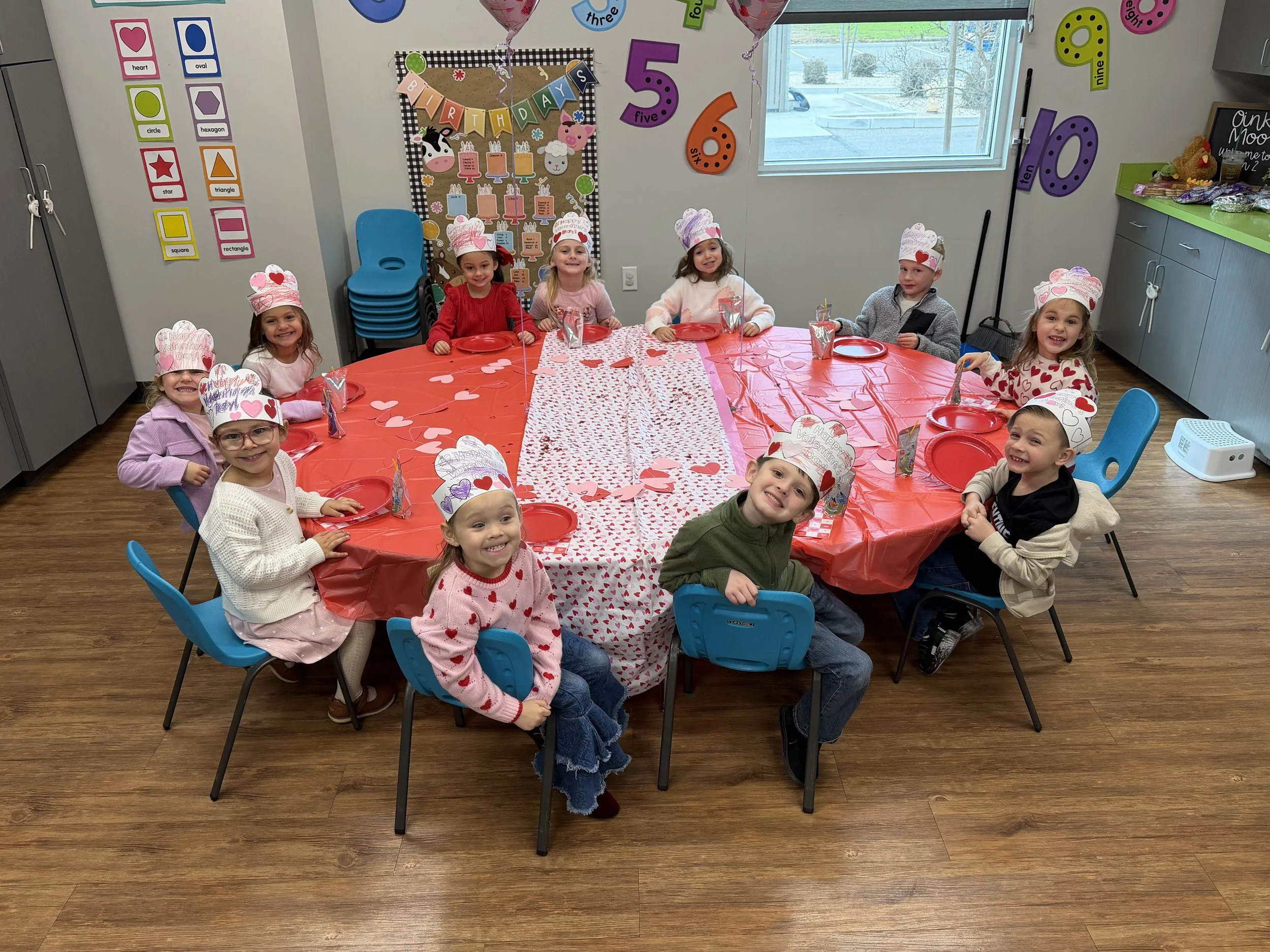 Children celebrating a birthday party, seated around a table with red, pink, and heart-themed decorations, wearing handmade paper hats with hearts, in a decorated classroom.
