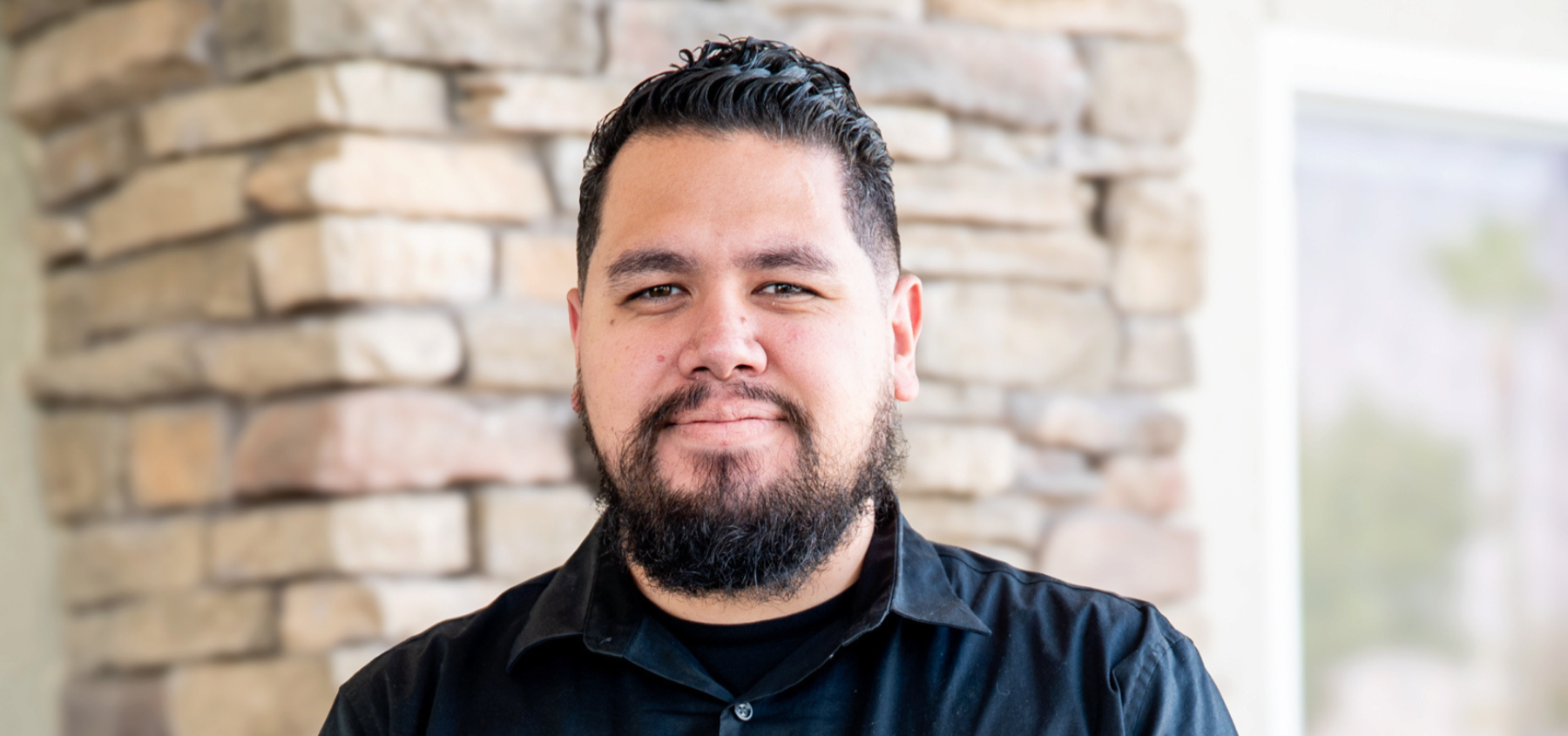 Portrait of a man with a beard and mustache, wearing a black shirt, smiling softly, standing in front of a stone wall and a large window with a blurred outdoor scene.