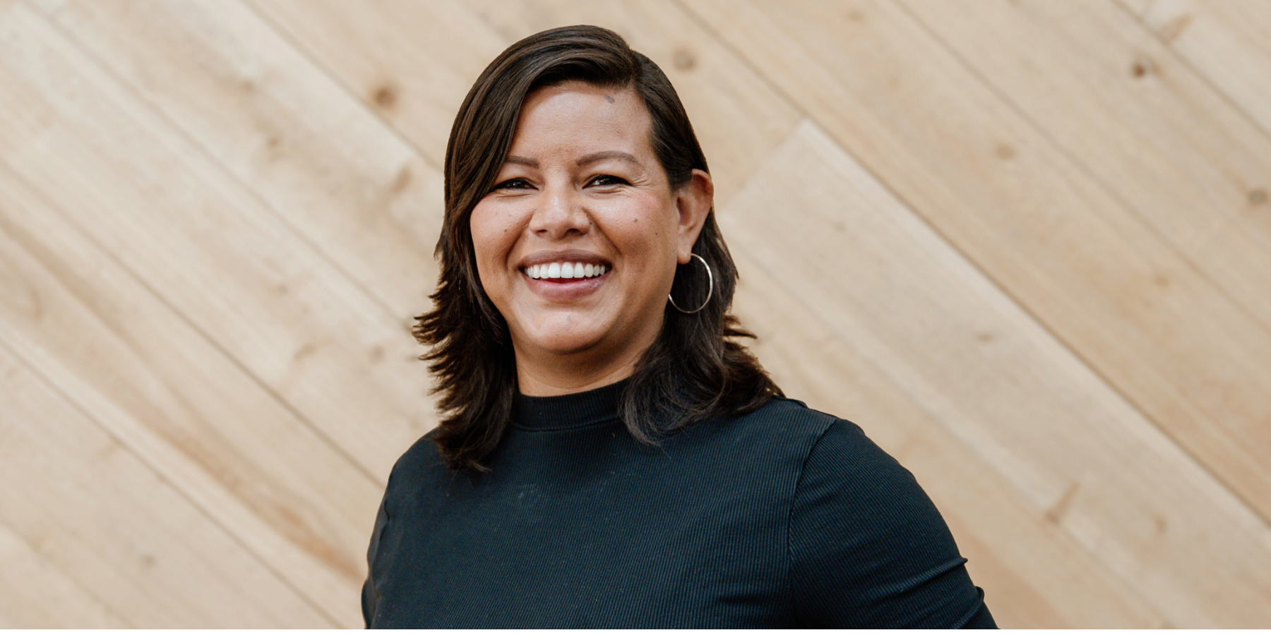 A smiling woman with dark brown hair, wearing hoop earrings and a black top, standing in front of a light wood-paneled wall.